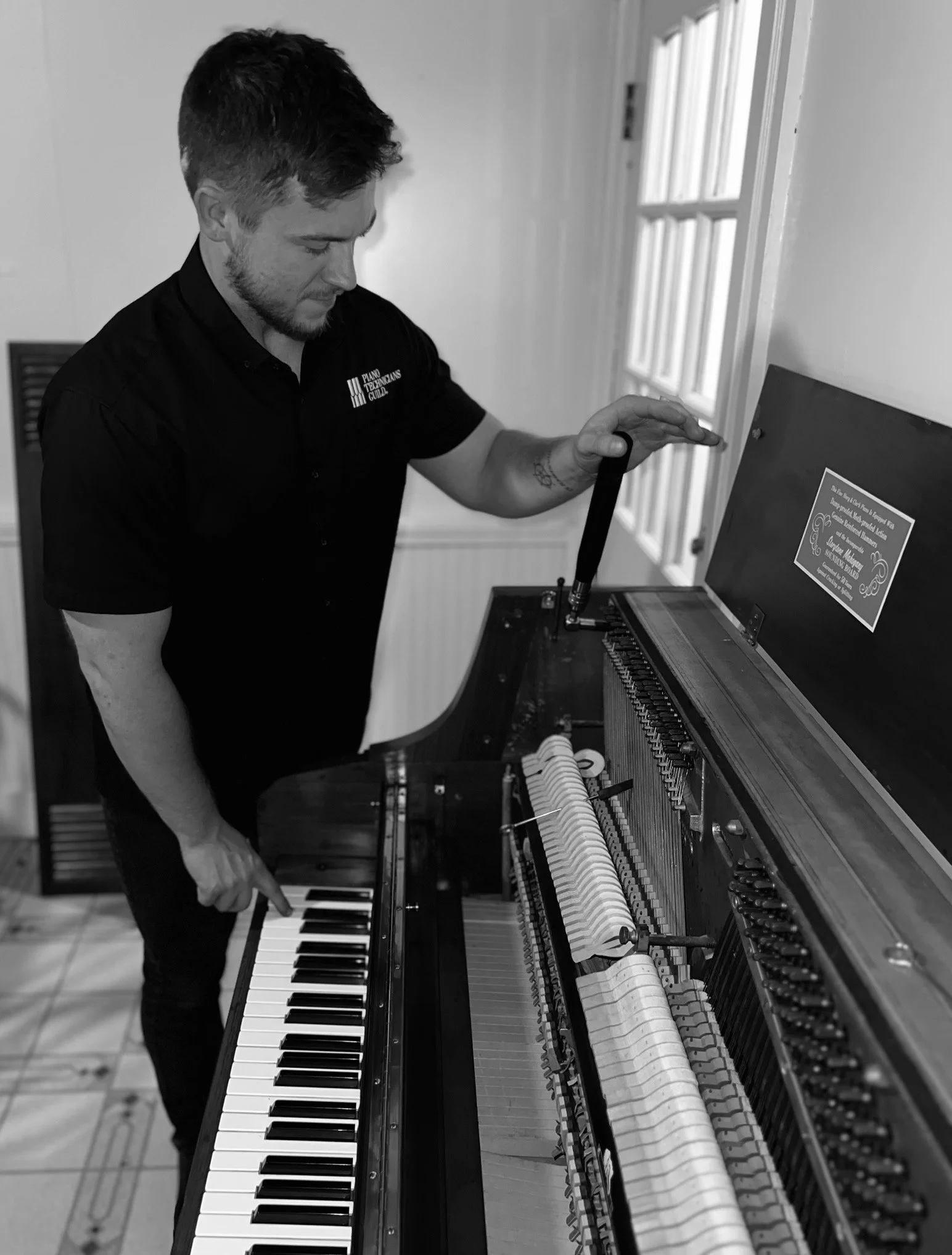 A man in a black shirt tuning an upright piano with a tuning hammer, opening the piano lid to view its strings and hammers.