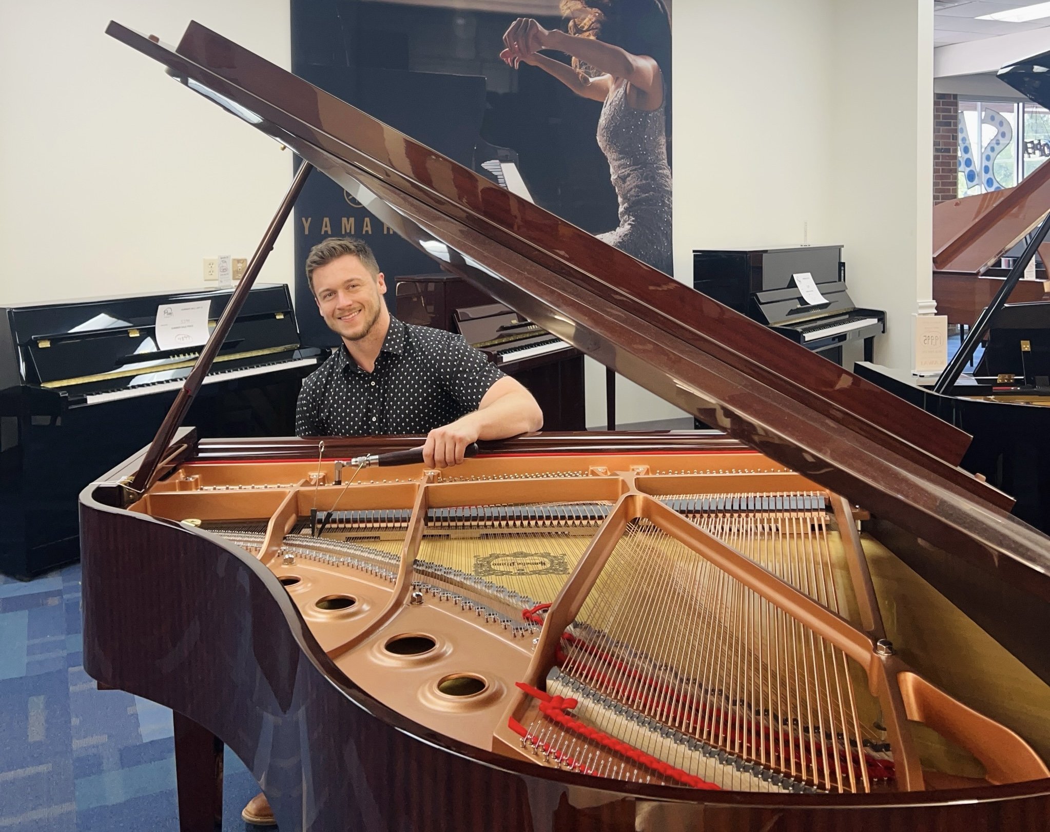 A smiling man sitting at a grand piano in a showroom, with other pianos visible in the background.