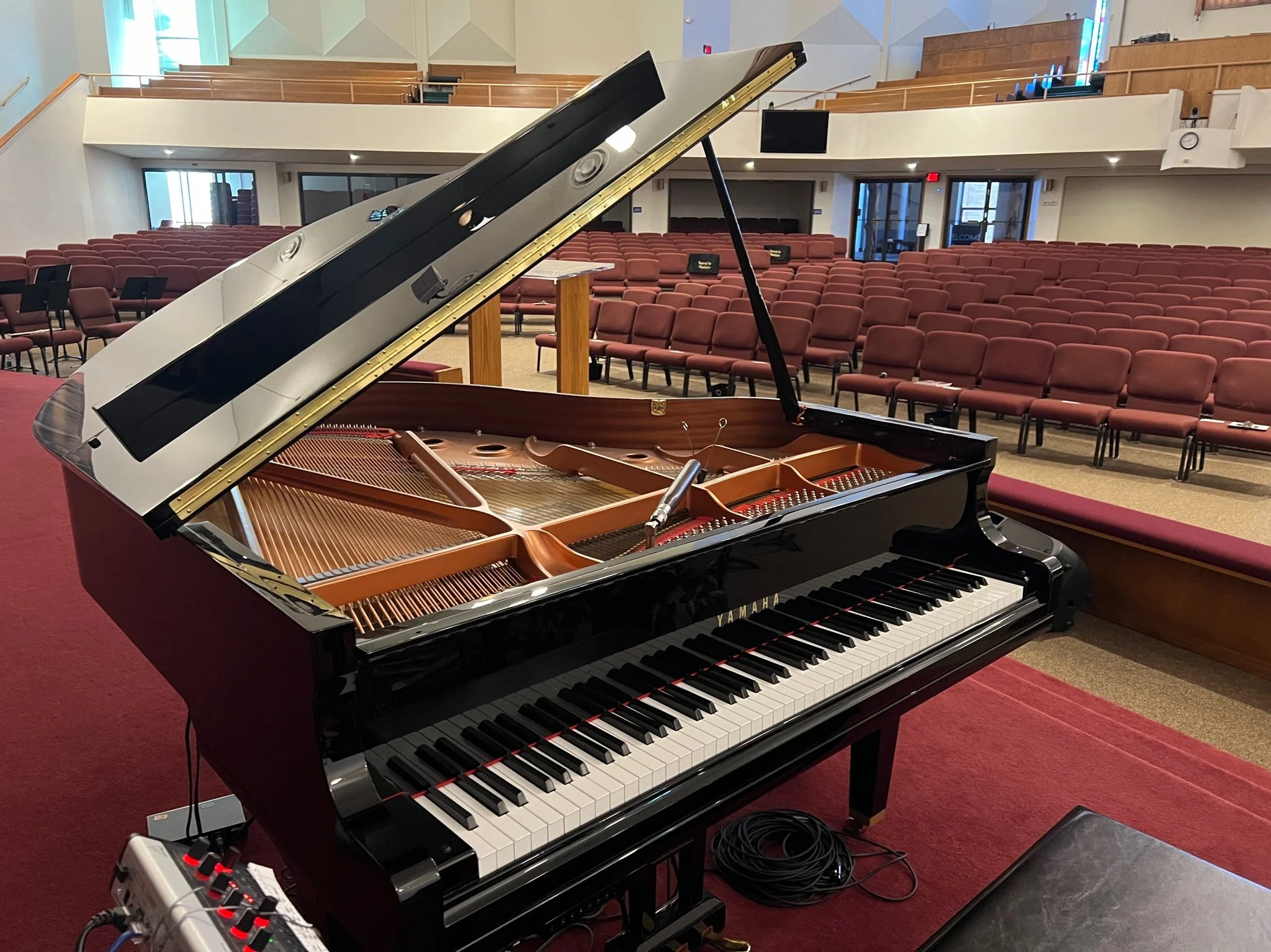A grand piano with its lid open inside a large auditorium with red chairs and a red carpet.