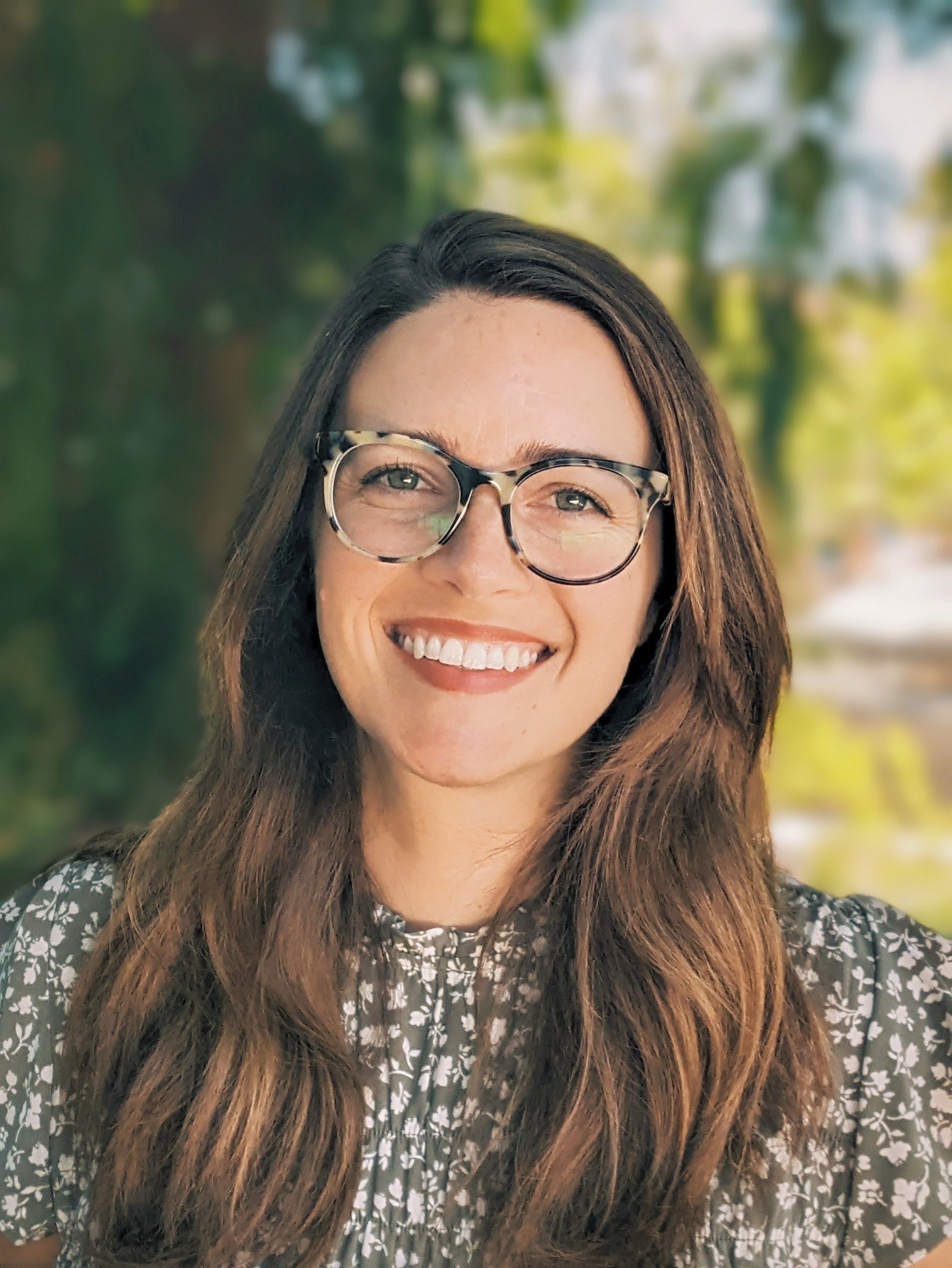 A young woman with long brown hair and glasses smiling outdoors, with blurred green trees and sunlight in the background.