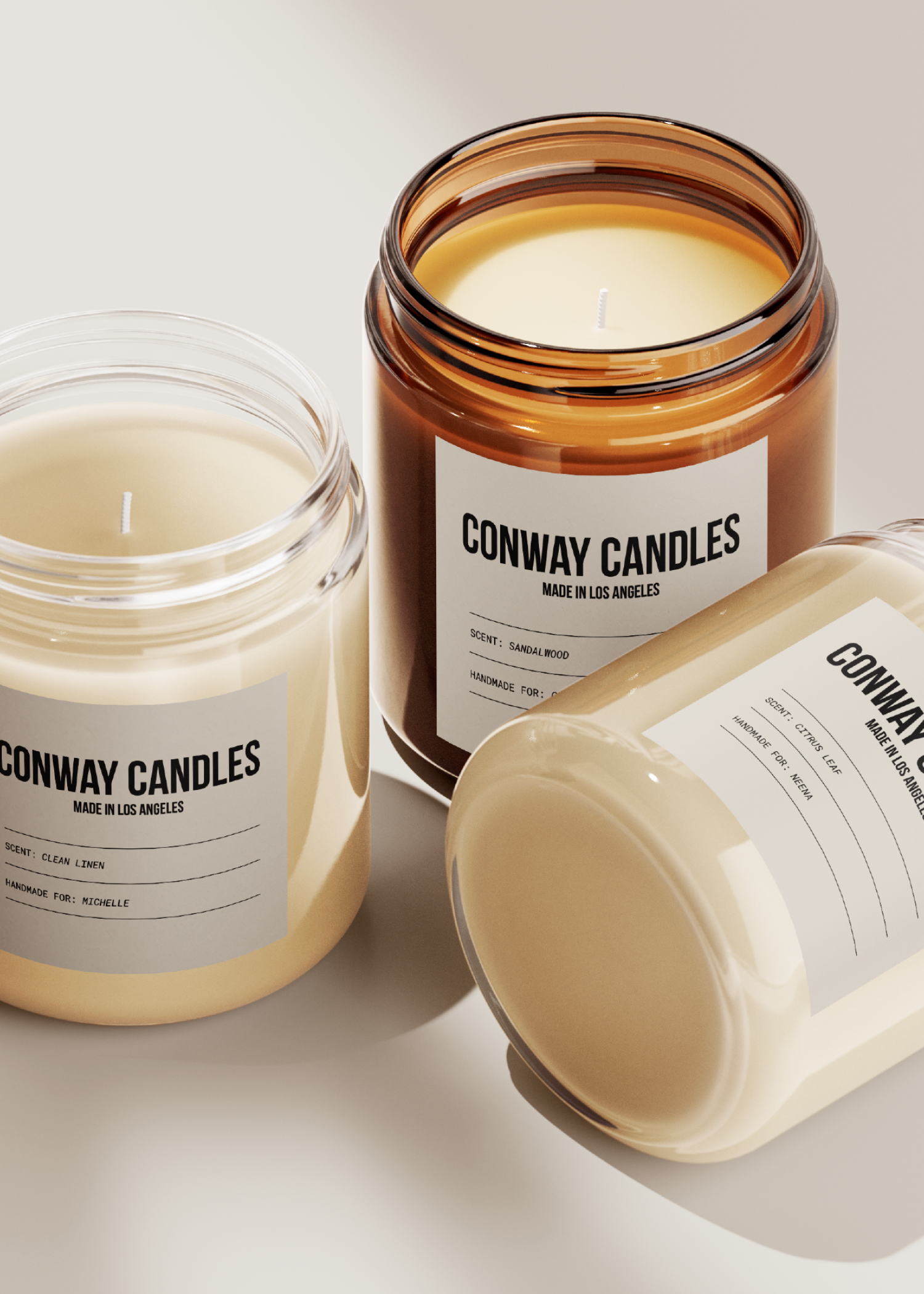 Three candles in glass jars with white labels, one amber-colored jar and two clear jars, all with unlit wicks, placed on a white surface.