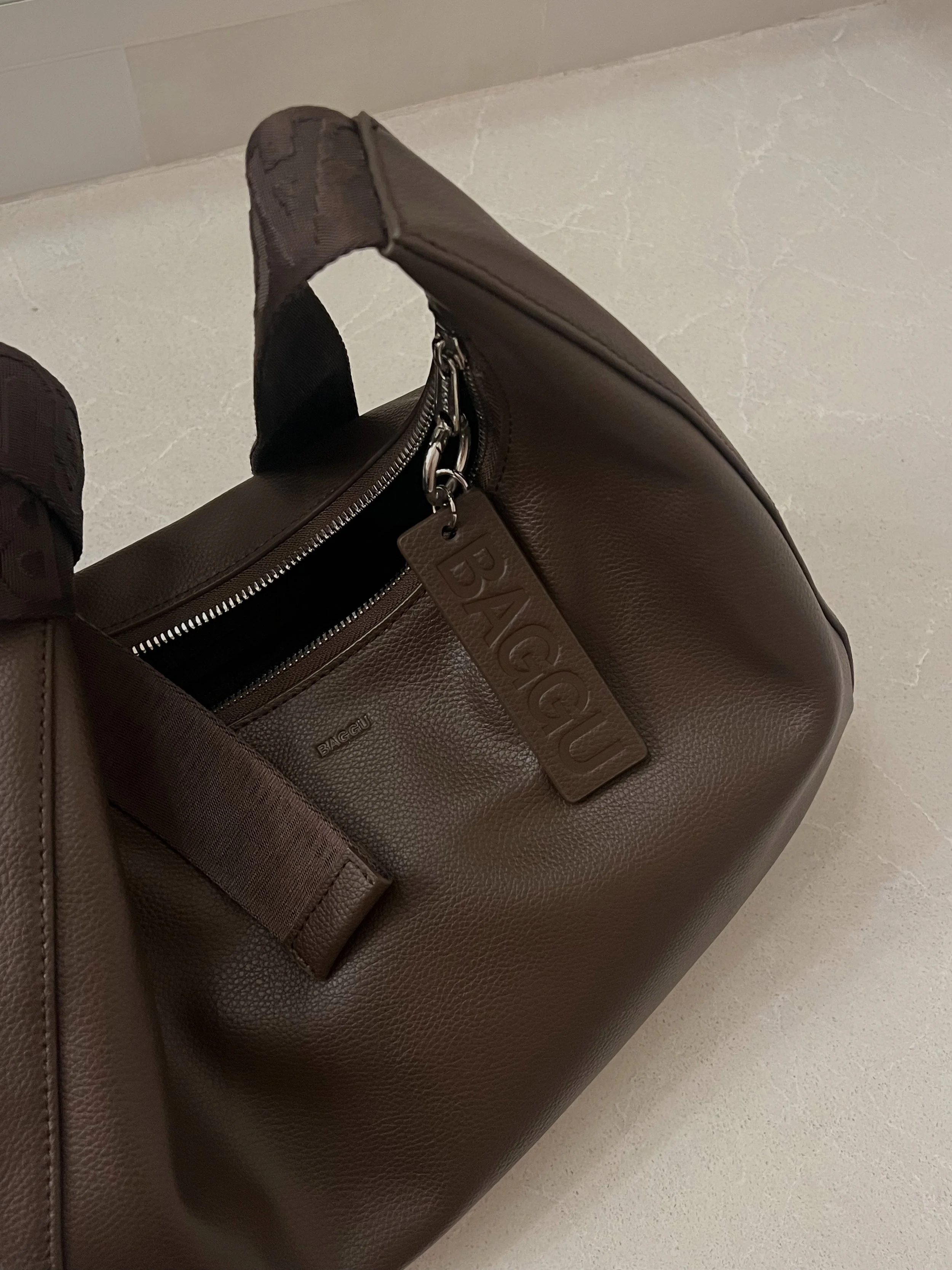A brown leather handbag with a handle, zipper, and a leather tag that reads 'BAGGU'