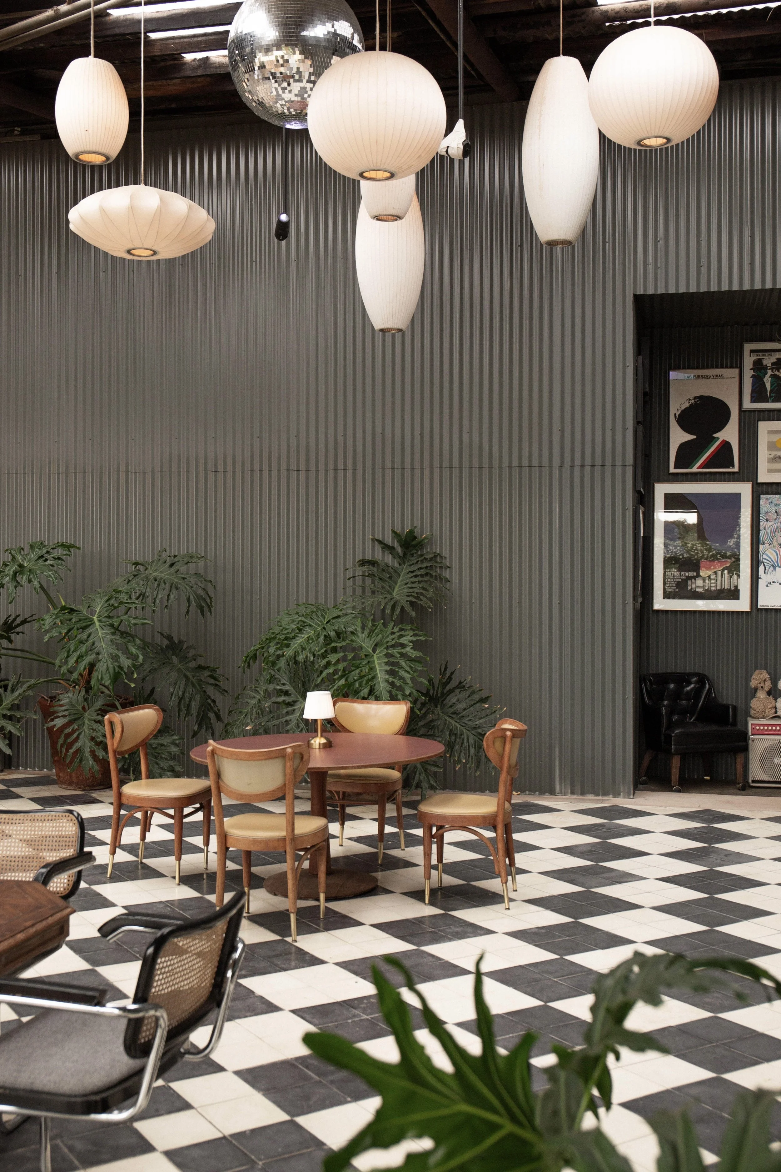 Interior of a stylish cafe or lounge with a checkered black and white floor, wooden chairs with beige cushions around a round table, and various hanging light fixtures including paper lanterns and a disco ball. Plants and framed artwork adorn the spa