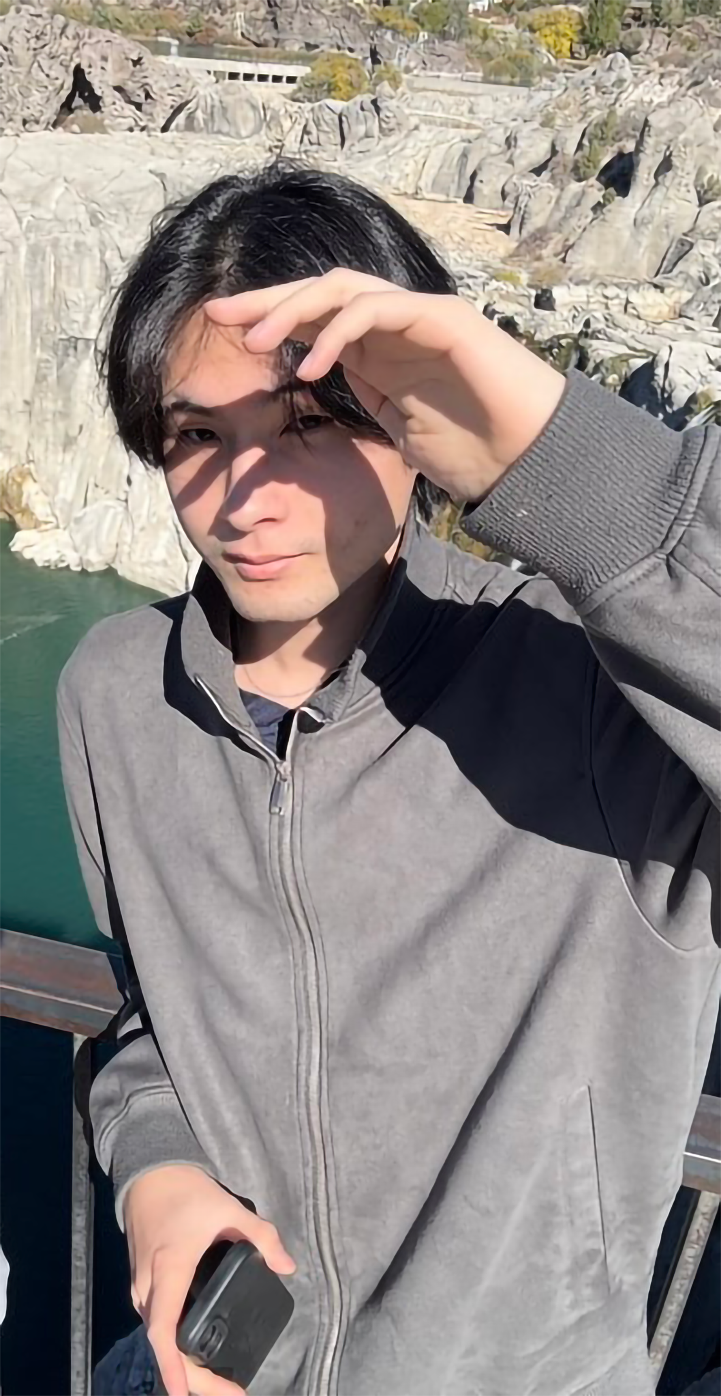 A young man with black hair, wearing a gray and black jacket, taking a selfie outdoors near a body of water with rocky cliffs in the background.