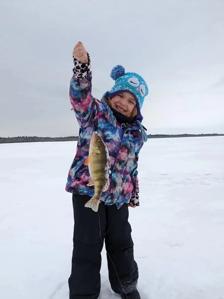 A young girl in a colorful winter jacket and a blue hat with a pom-pom, standing on a snowy ice-covered landscape, holding up a fish she caught.