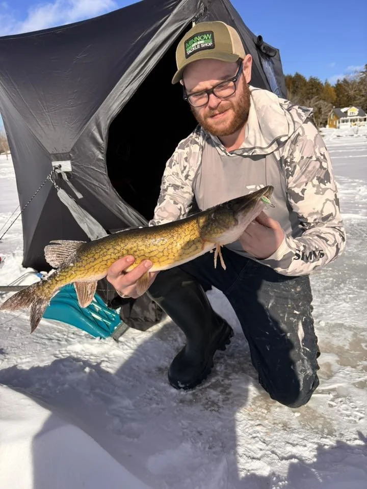 A man in winter clothing and a cap kneels on a snow-covered ice surface, holding a large fish he just caught. He is close to a black ice fishing tent with a zippered entry. The background shows snow-covered ground and some houses with trees.
