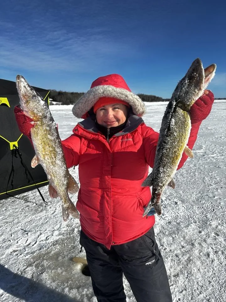 Child in red winter coat and Santa hat holding two fish on a snowy frozen lake.