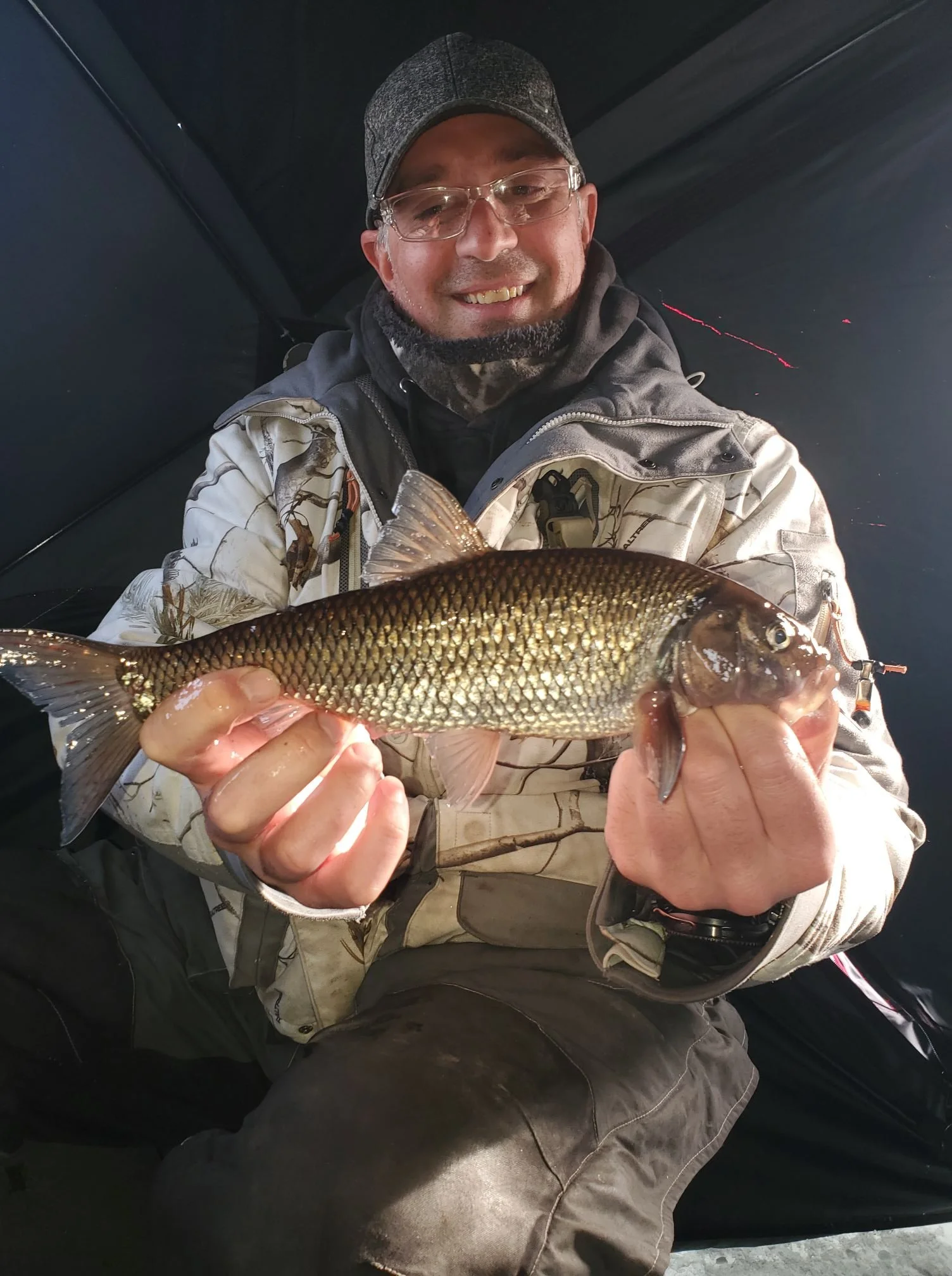 Man in outdoor clothing smiling and holding a fish inside a dark shelter or tent.
