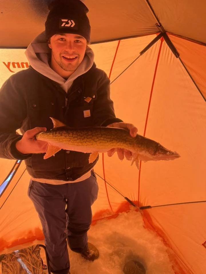 A man inside an orange ice fishing tent holding a large fish, smiling at the camera.