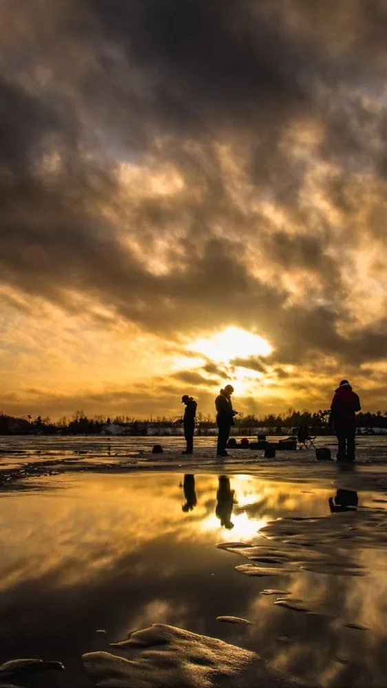 Three people ice fishing on a frozen lake with a dramatic sunset sky and their reflections in the water.