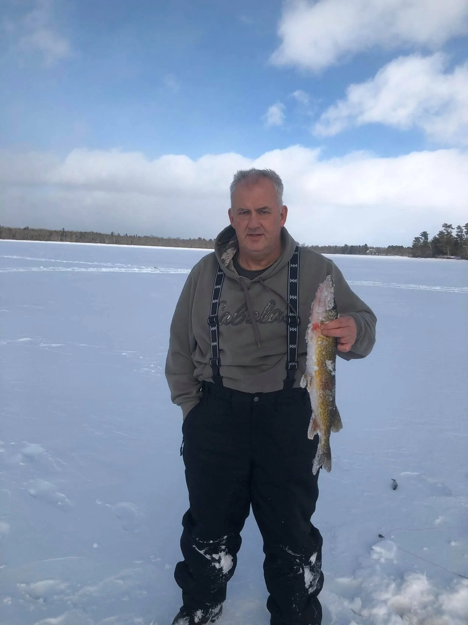 Man standing on snow-covered ice holding a large fish, with a winter landscape and cloudy sky in the background.