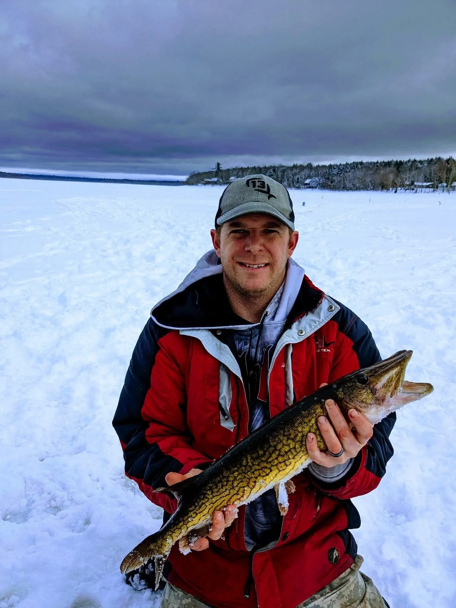 A man in winter clothing holding a large fish on a snowy landscape with a frozen lake and trees in the background.