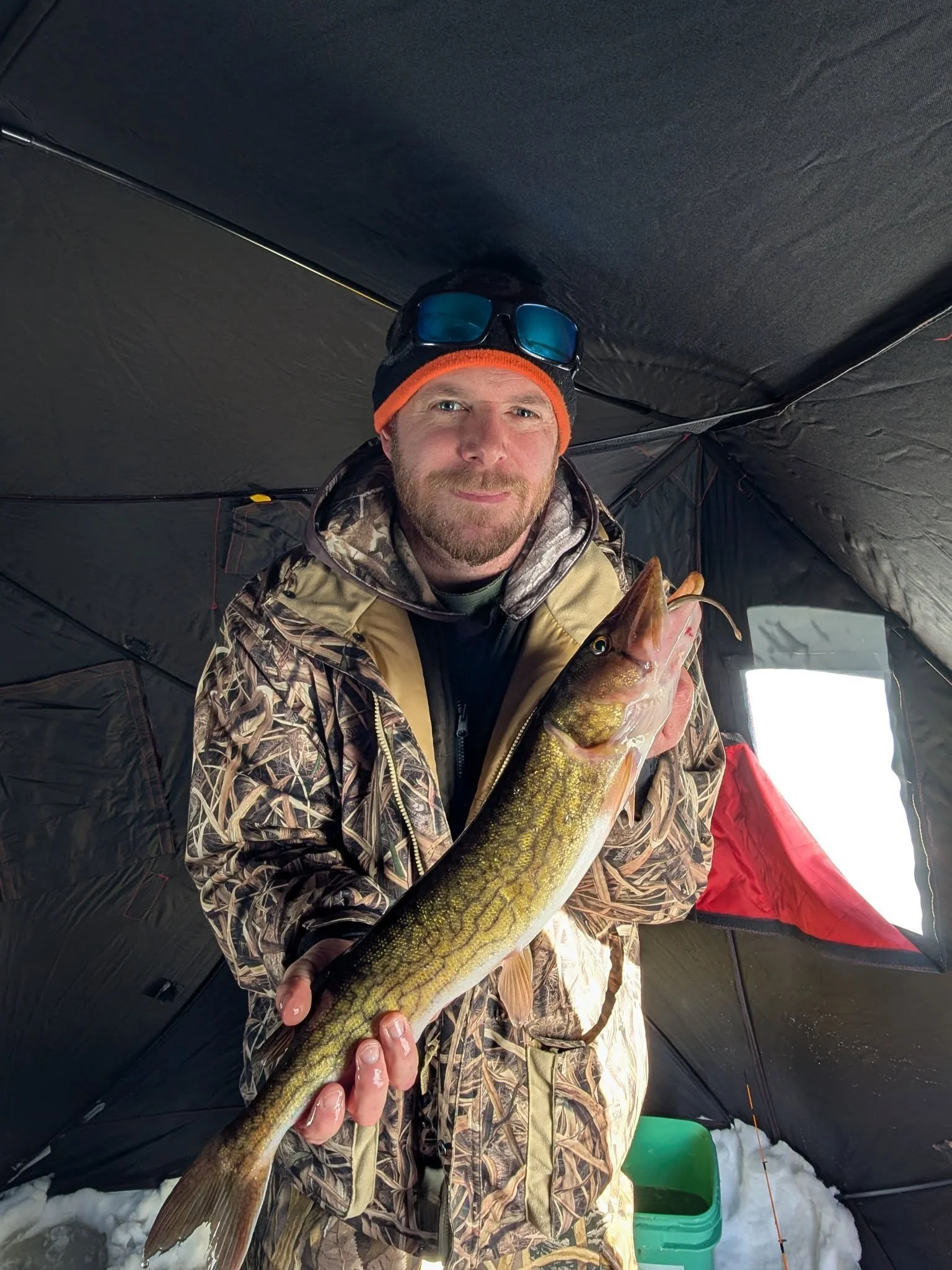 Man in camouflage winter gear holding a large fish, standing inside an ice fishing shelter with snow on the ground.