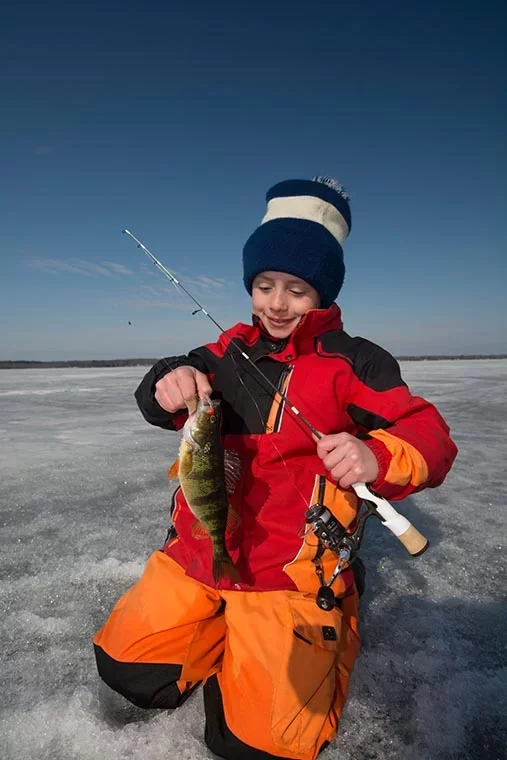 A boy in winter clothing kneels on ice while holding a fishing rod and a freshly caught fish, with a clear blue sky in the background.