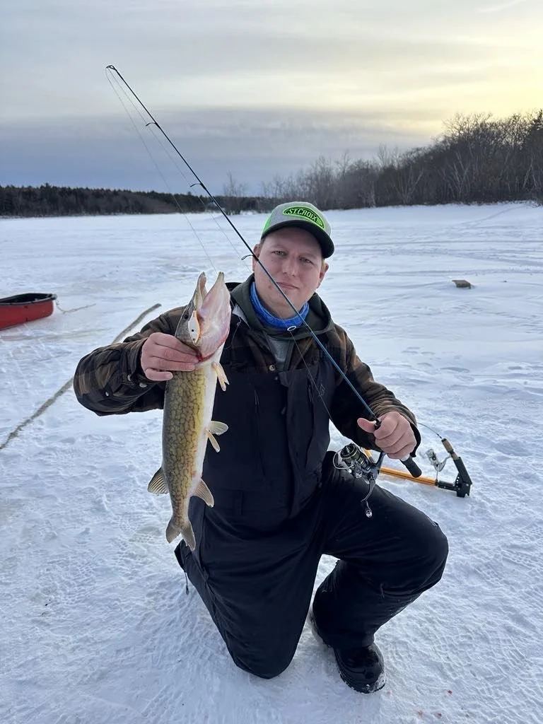Man kneeling on ice holding a fish with a fishing rod in a snowy landscape.