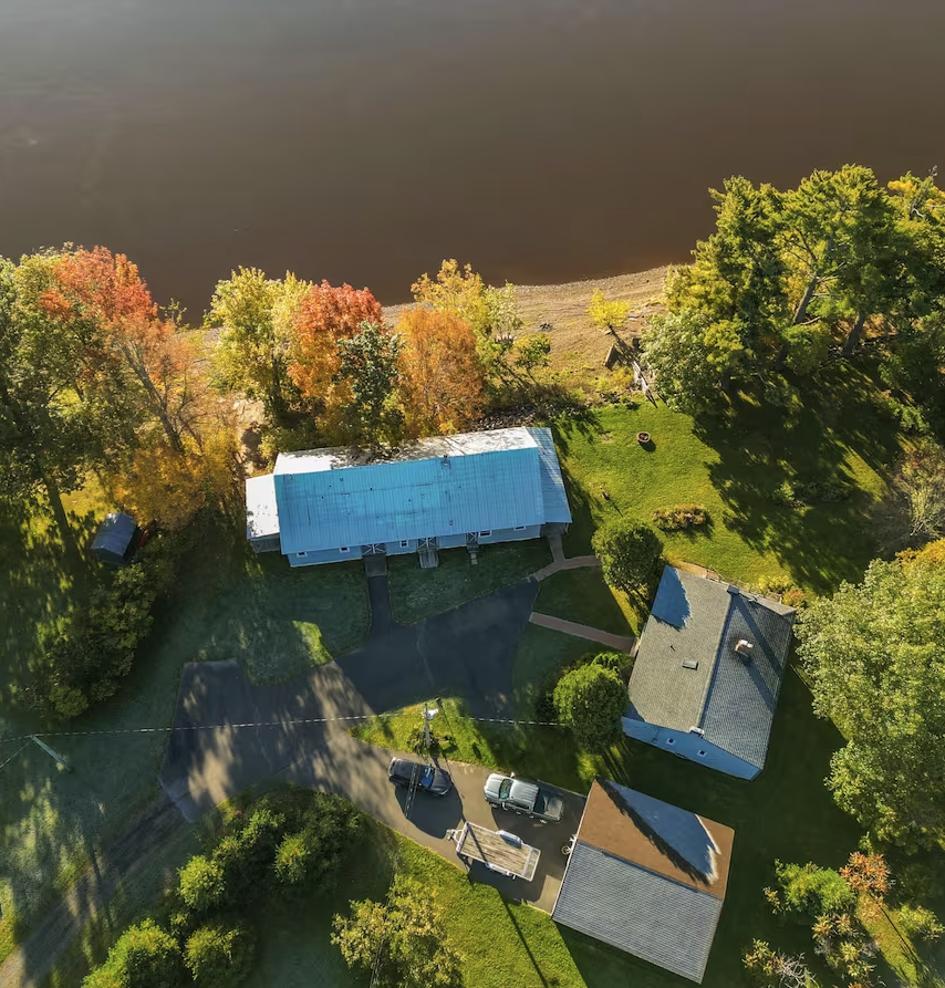 An aerial view of a property near a river, featuring several buildings, trees with autumn foliage, a driveway with parked cars, and a grassy yard.