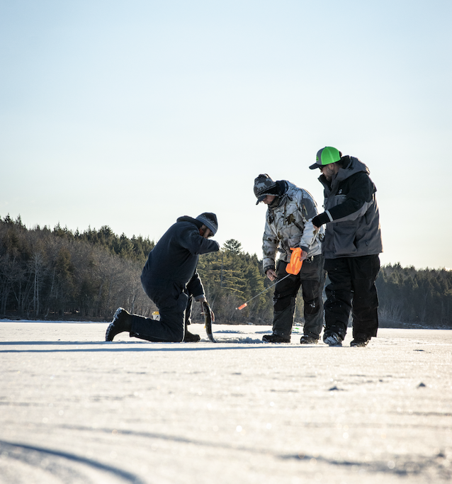 Three people ice fishing on a frozen lake, with a forest in the background, wearing winter clothing and holding fishing equipment.