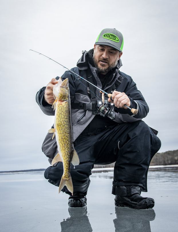 Man wearing a gray and green cap kneeling on icy water, holding a fishing rod in one hand and a yellowish fish in the other.