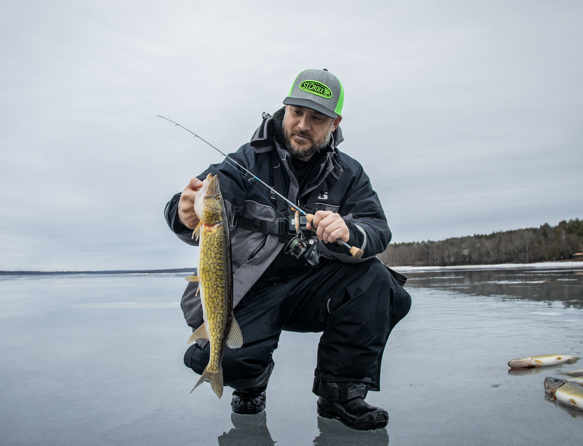Man kneeling by the edge of a lake, holding a fishing rod and a large fish he caught.