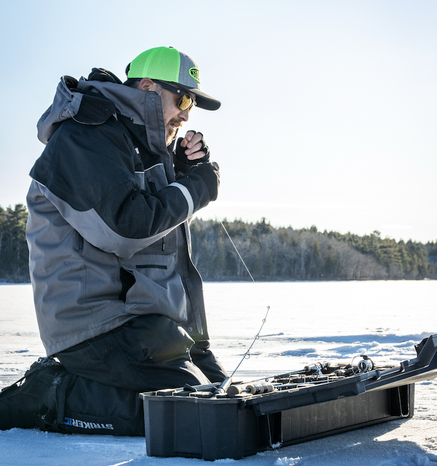 Man in winter clothing ice fishing on frozen lake, sitting next to a tackle box with fishing rods, wearing a neon green cap, sunglasses, and gloves.