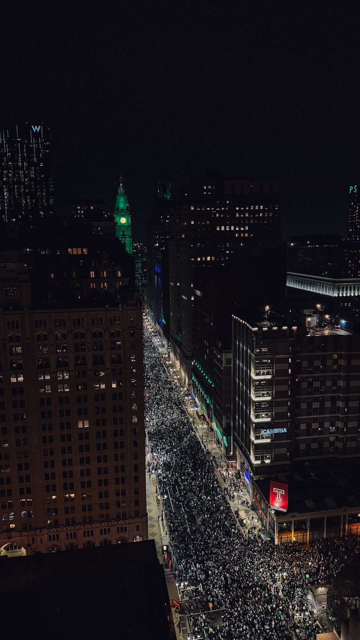 Nighttime city street filled with people, high-rise buildings, illuminated signs, and a clock tower in the distance.