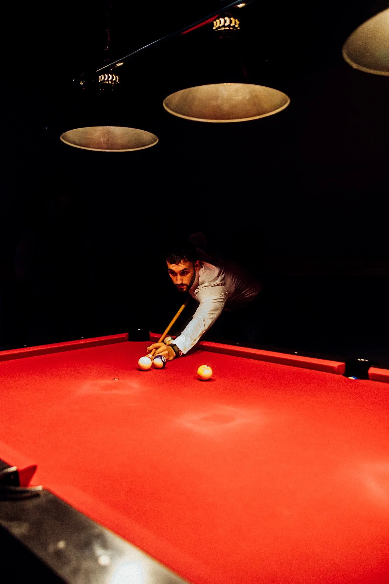 A man in a white shirt playing pool on a red billiard table in a dimly lit room.