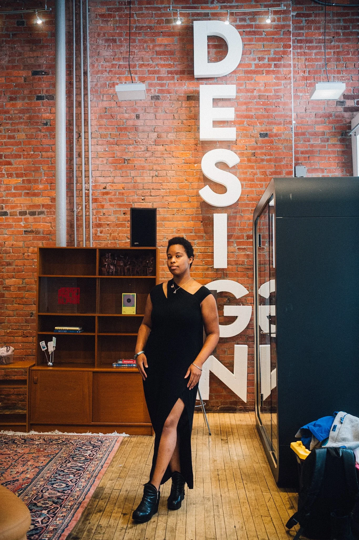 A woman in a black dress with a high slit standing in front of a brick wall with large white letters spelling 'DESIGN' vertically.
