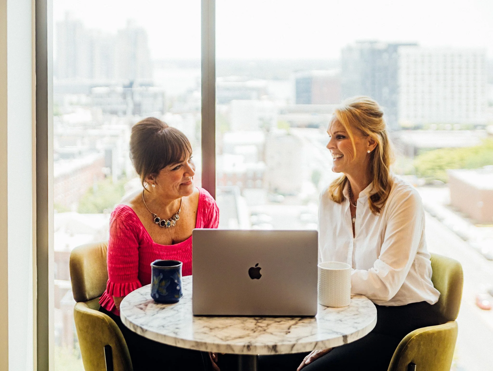 Two women sitting at a round marble table with a laptop and coffee mugs, smiling and talking in a high-rise office with large windows and city view.