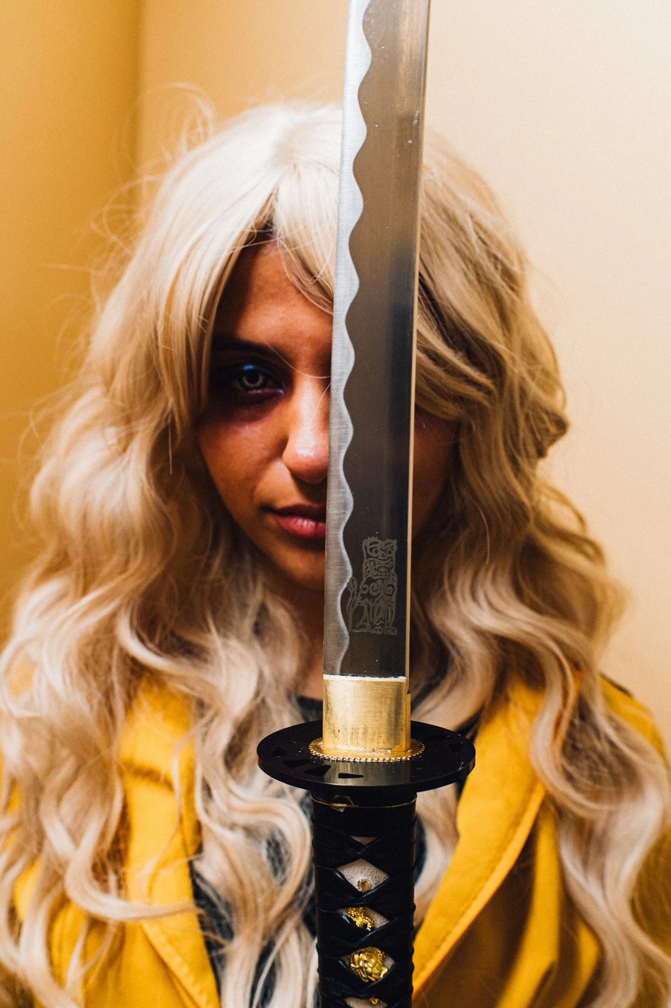 A person with long, wavy blonde hair holding a sword vertically in front of their face, half obscuring their face, with dark eye makeup on one eye and wearing a yellow jacket.