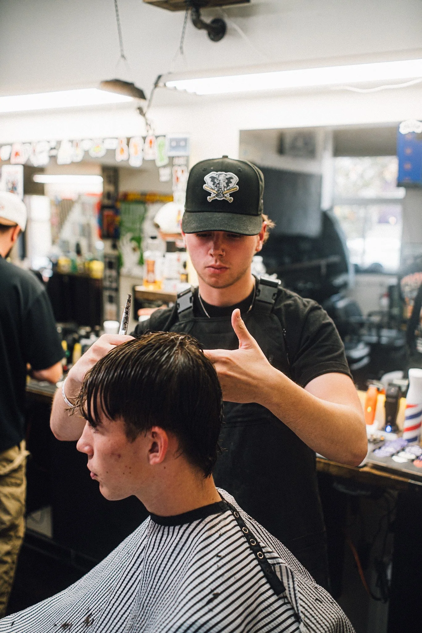Young man getting a haircut at a barbershop, barber is wearing a black cap and black t-shirt, focused on cutting client's hair.