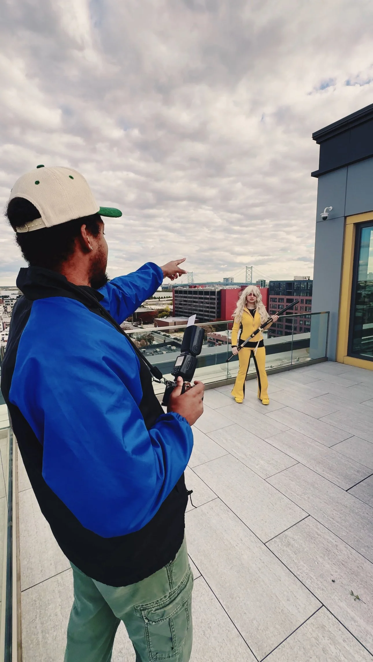 A man in a blue jacket and beige cap is pointing at a woman dressed in a yellow costume on a rooftop, with a cityscape and a bridge in the background.