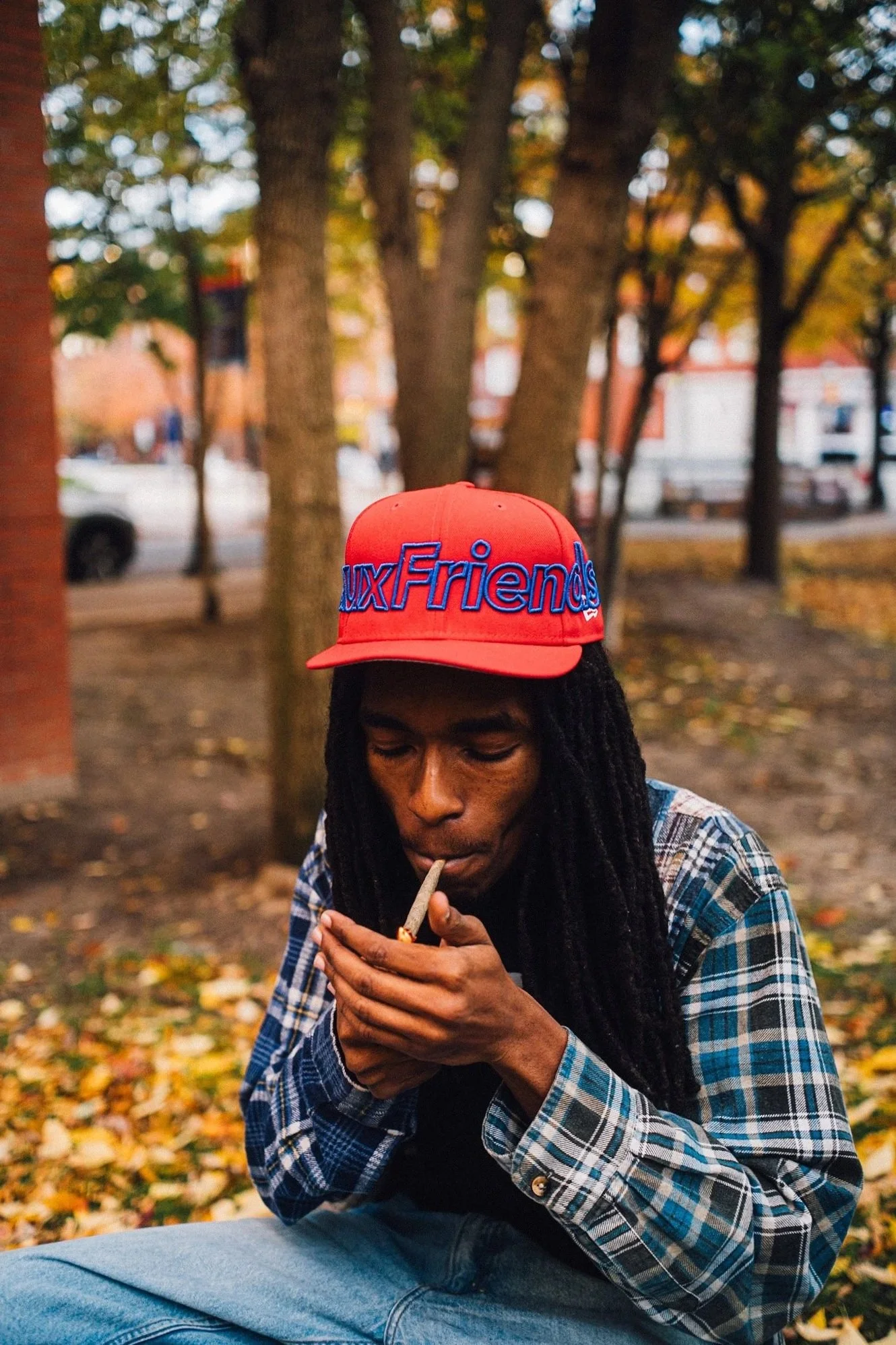 A man with long dark dreadlocks wearing a red cap with blue embroidery, a plaid shirt, and jeans, sitting outdoors in a park with autumn leaves on the ground, lighting a cigarette.