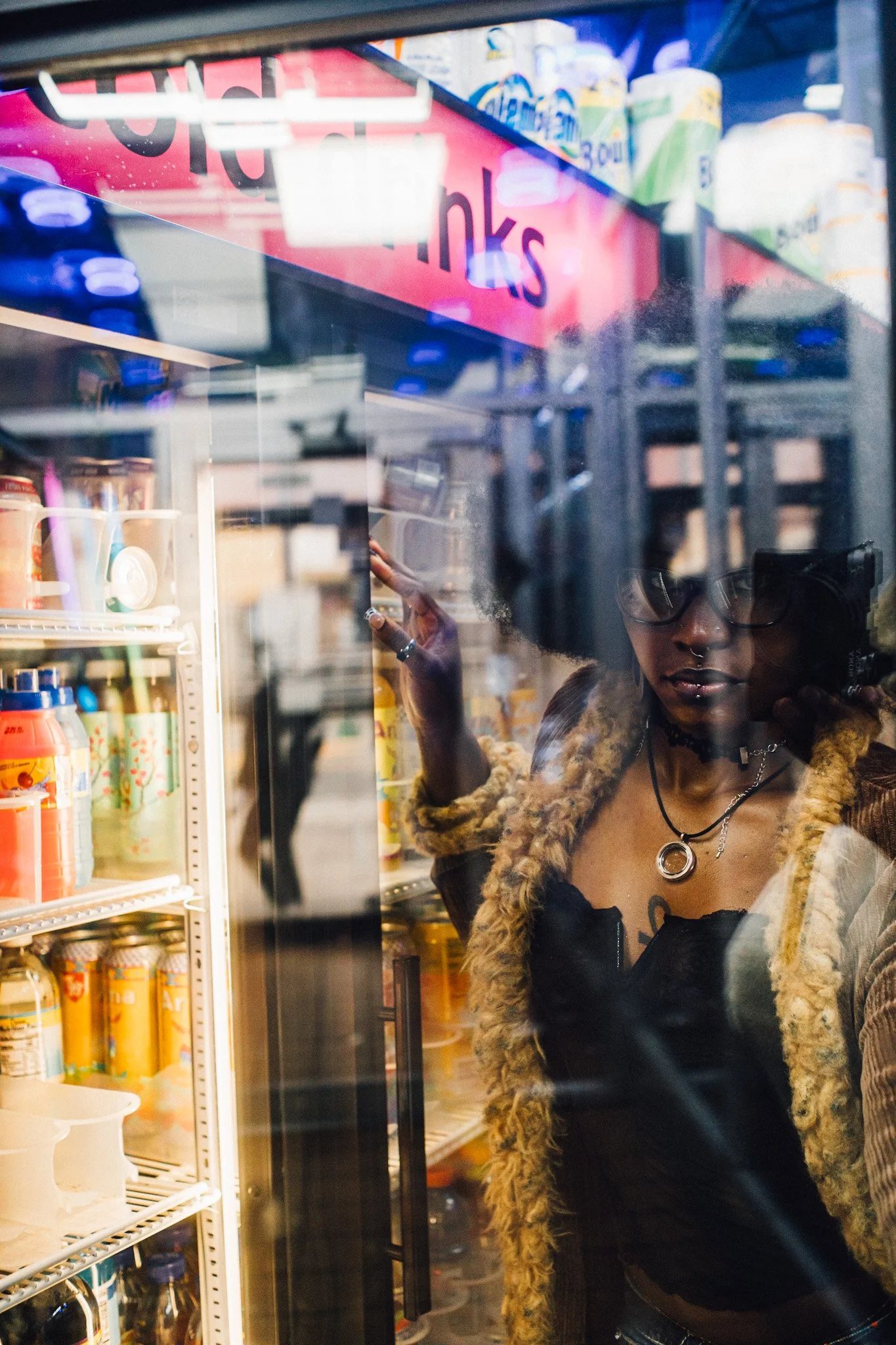 A woman with curly hair and glasses looking at a refrigerator in a store, with shelves of drinks and snacks visible.