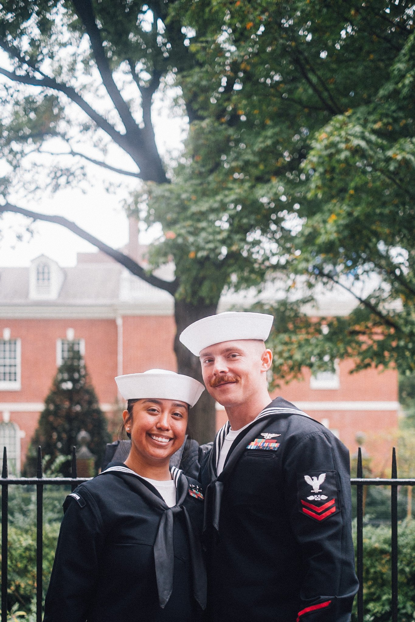 Two sailors in uniform, a woman and a man, smiling and standing close together outdoors with trees and a brick building in the background.
