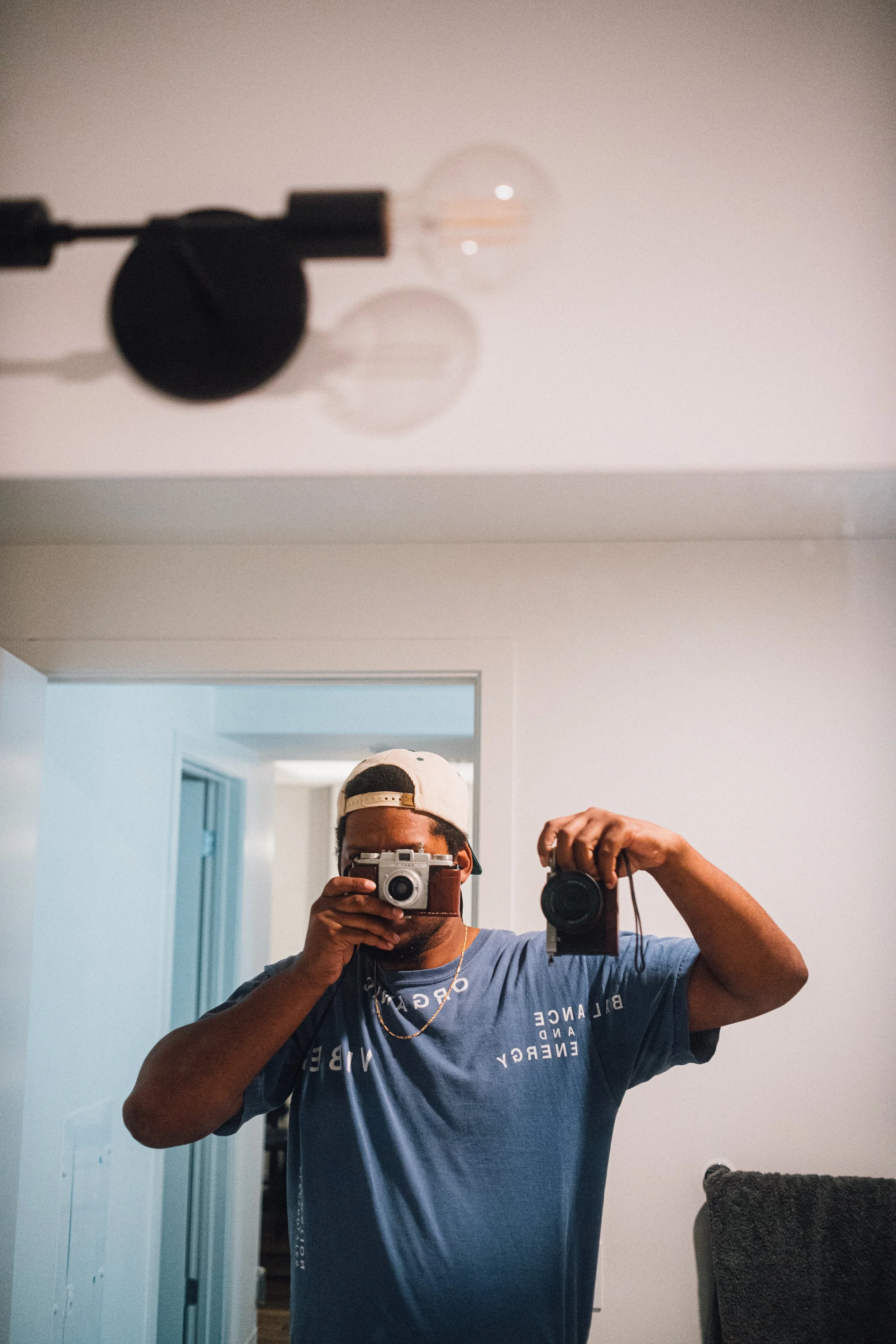 A person taking a mirror selfie with a camera, wearing a blue t-shirt and a white cap turned backward in a room with minimal decor.