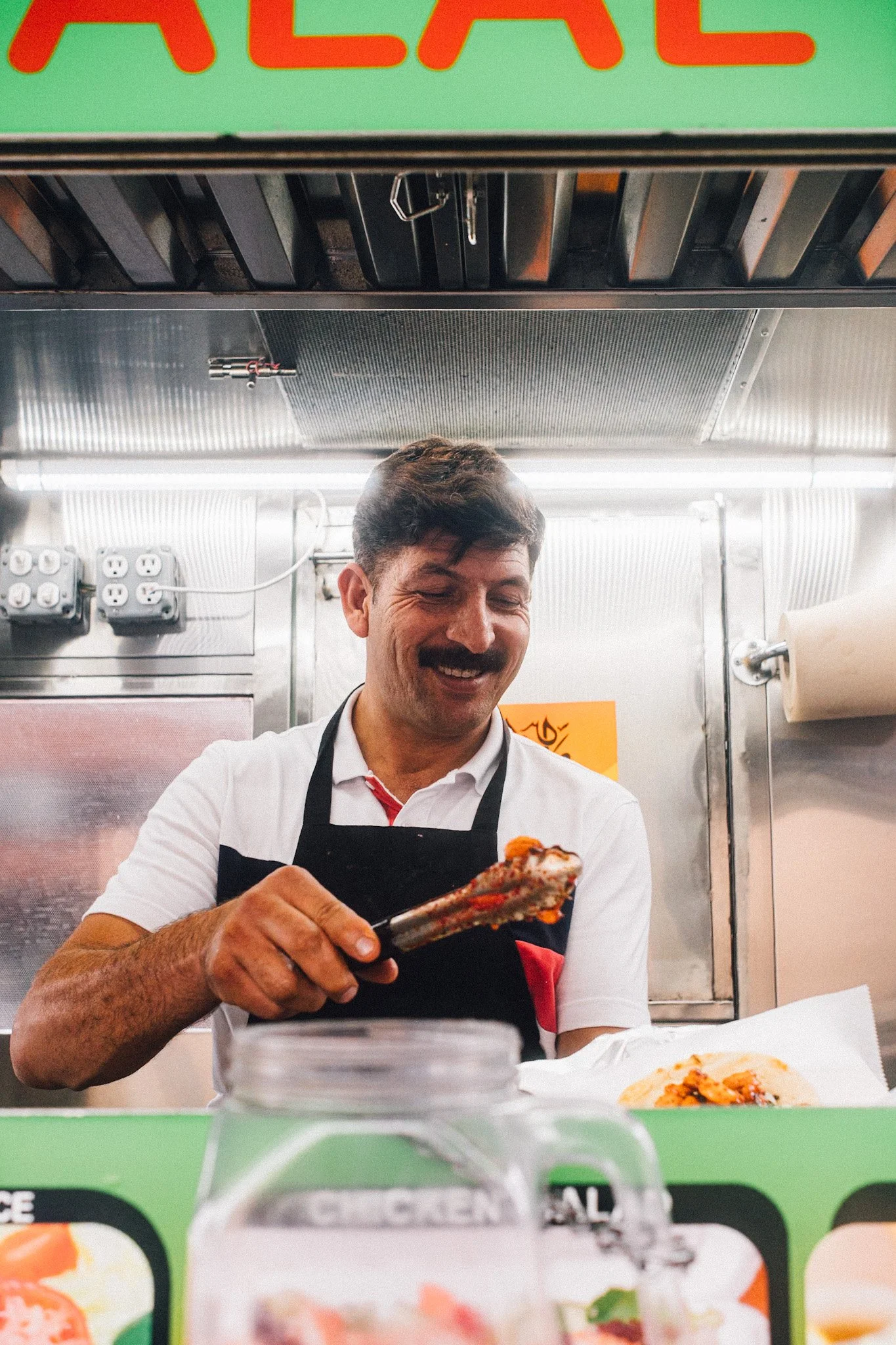A smiling man in a white shirt and black apron preparing food at a food stand, holding tongs with a piece of meat.
