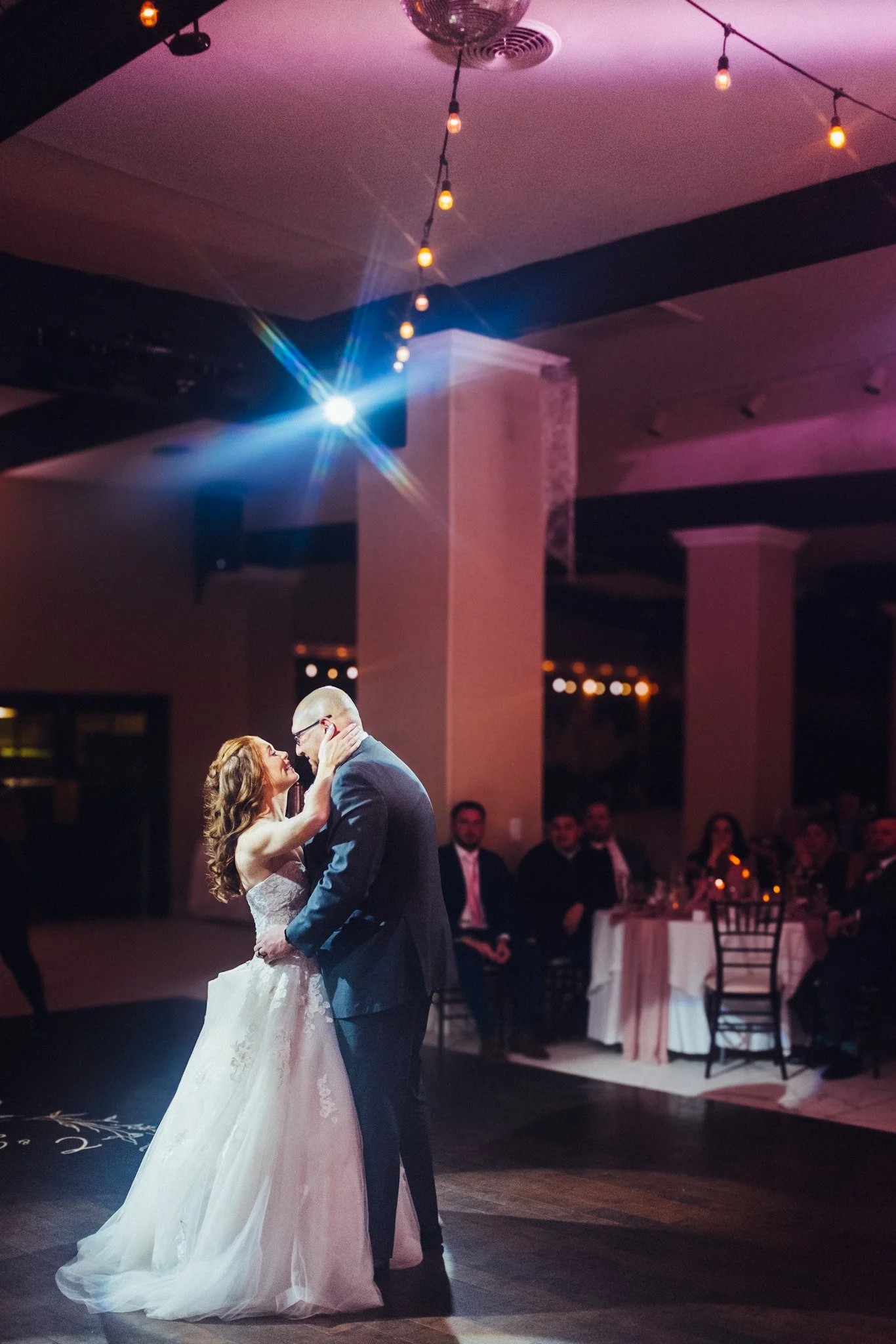 A bride and groom sharing their first dance at their wedding reception, with guests seated at tables in the background.