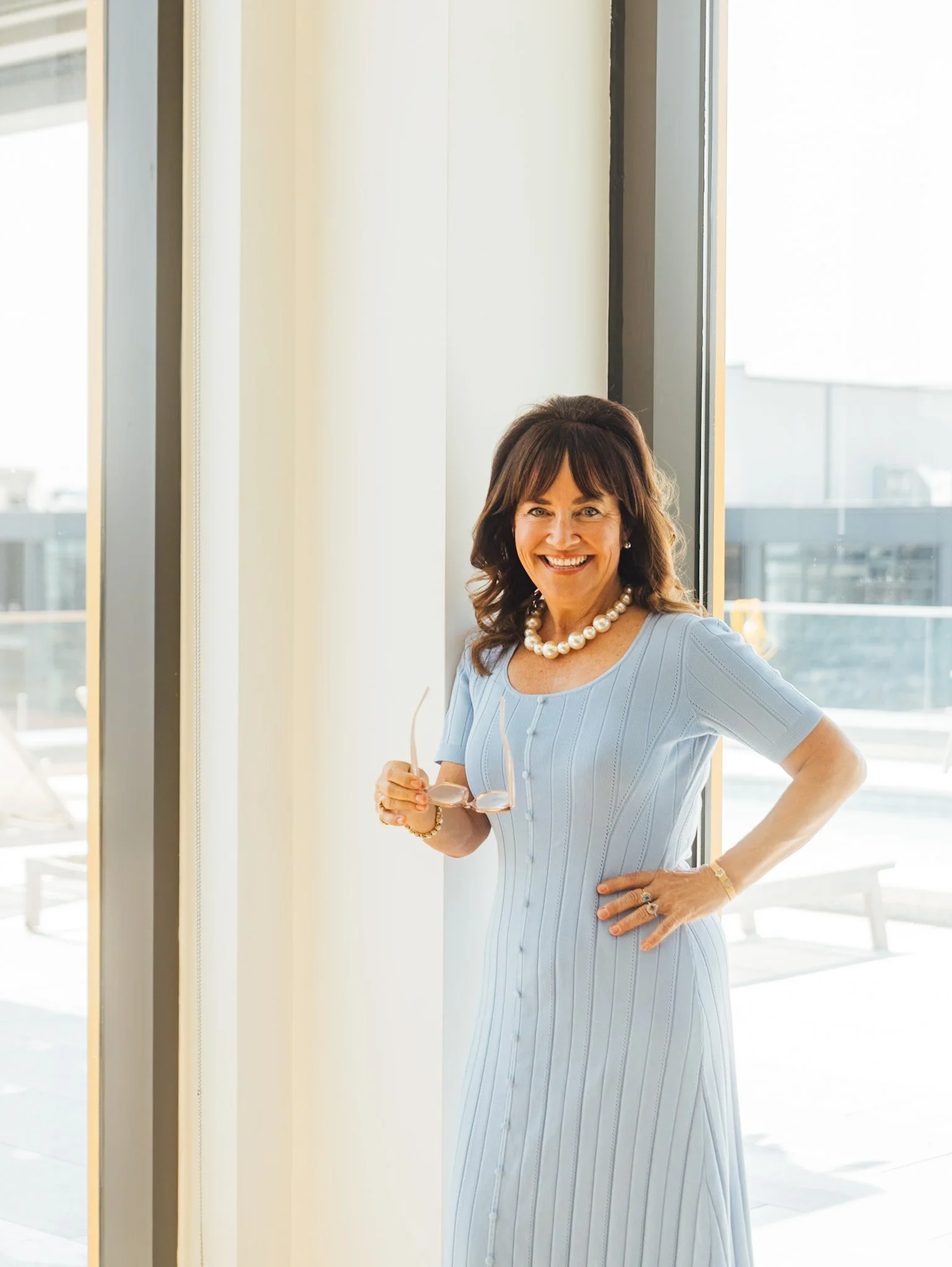 Smiling woman in a light blue dress and pearl necklace holding sunglasses stands next to large window in a bright room.