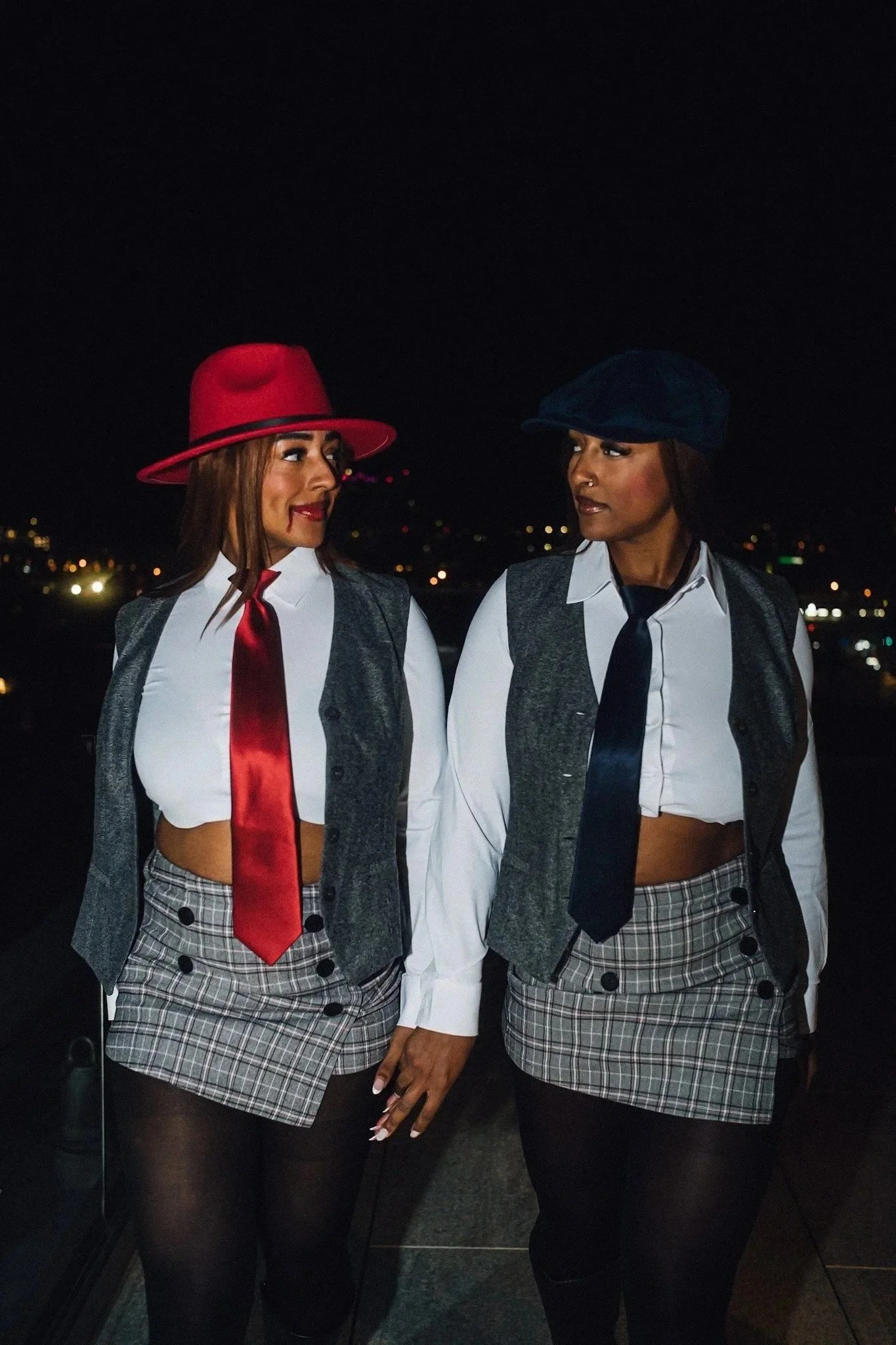 Two women dressed in schoolgirl costumes holding hands at night, with cityscape lights in the background.
