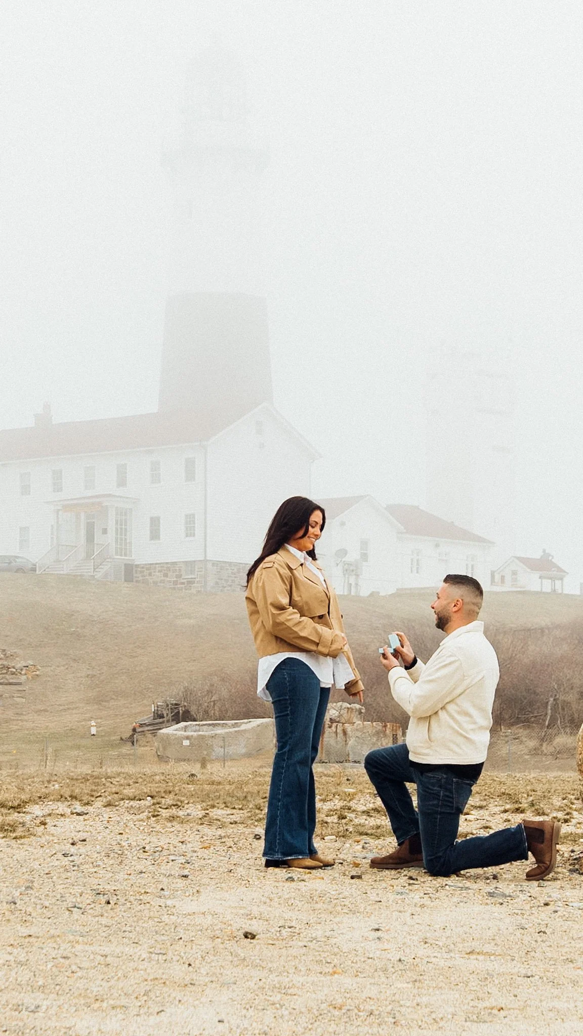 A man kneeling on one knee proposing to a woman outdoors on a foggy day, with a house in the background.