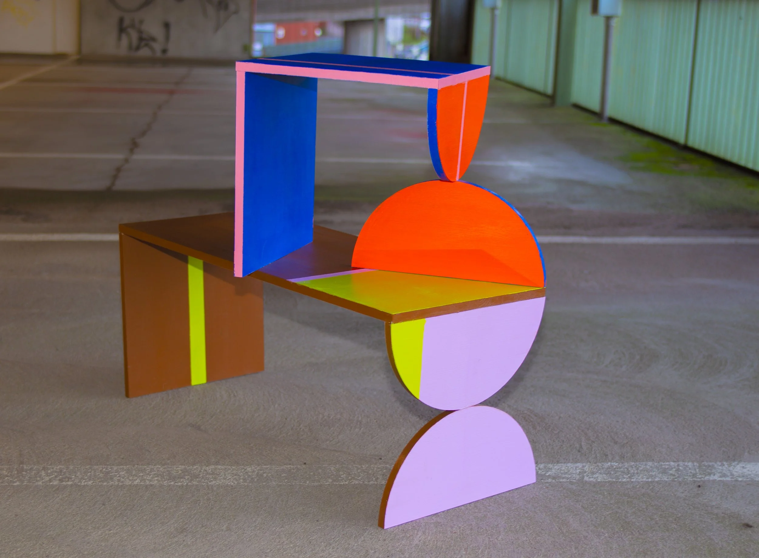 Värikkäitä geometrisia muotoja ja särmiöitä muodostava taideteos, joka koostuu puulevyistä, jotka on maalattu erivärisiksi ja aseteltu tasaiselle betoniparkkeerausalueelle.