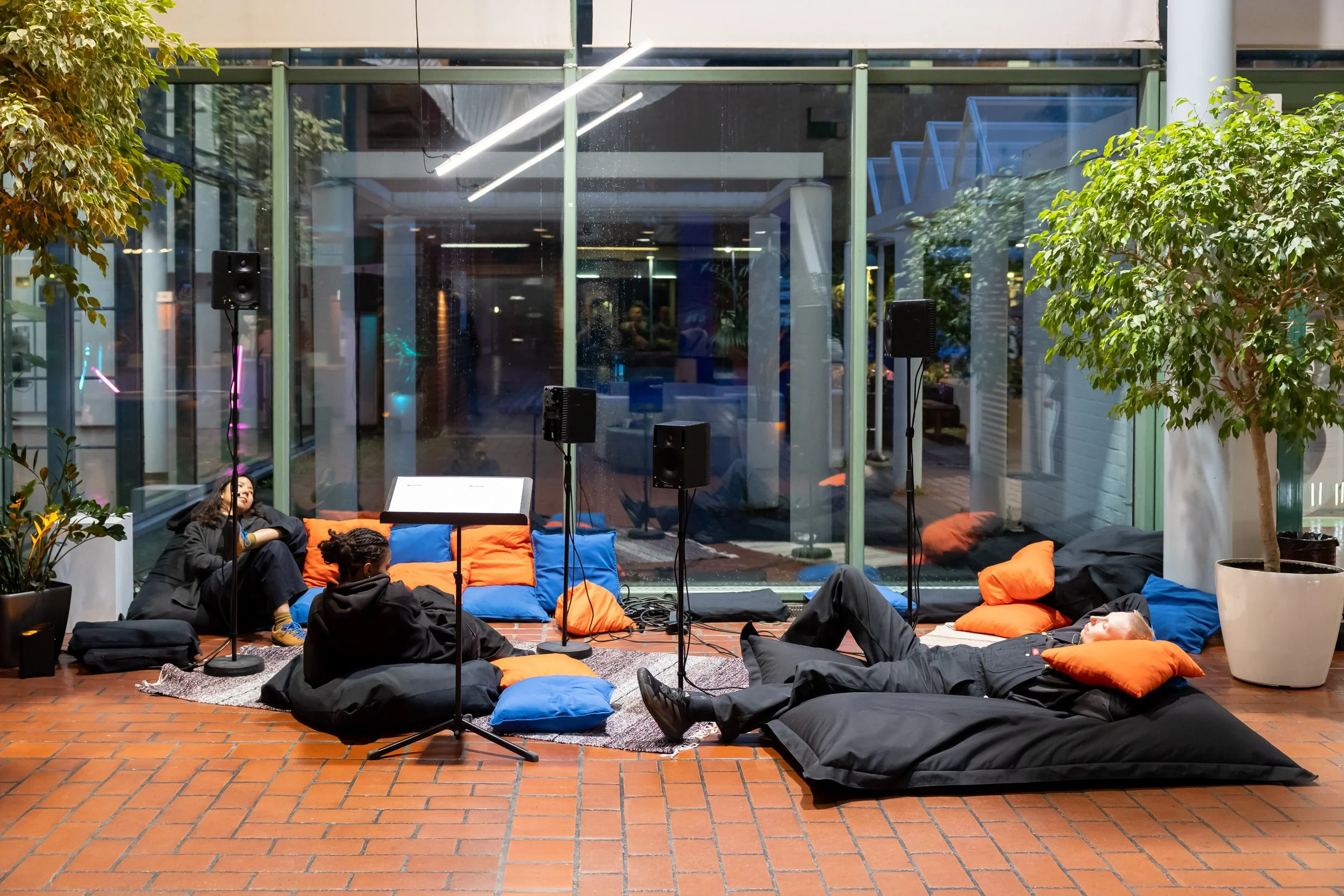 Kolm ihmistä makaa ja istuu pehmeillä täytetyillä lattialla ja sohvilla, ympärillä salkku, mikrofonit, kaiuttimet ja sisäkasveja, lasiseinän taustalla näkyy rakennuksen sisätiloja.