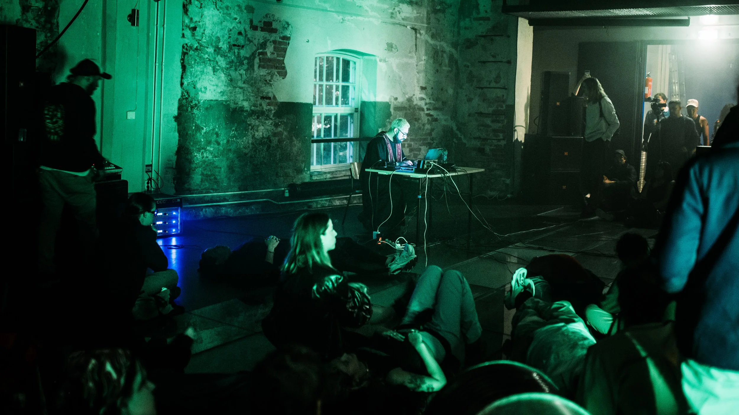 Valoton musiikkilava, jossa dj soittaa musiikkia vanhan kiviseinän edessä. Yleisö istuu ja makaa lattialla, valaistus on vihreä ja tunnelmallinen.