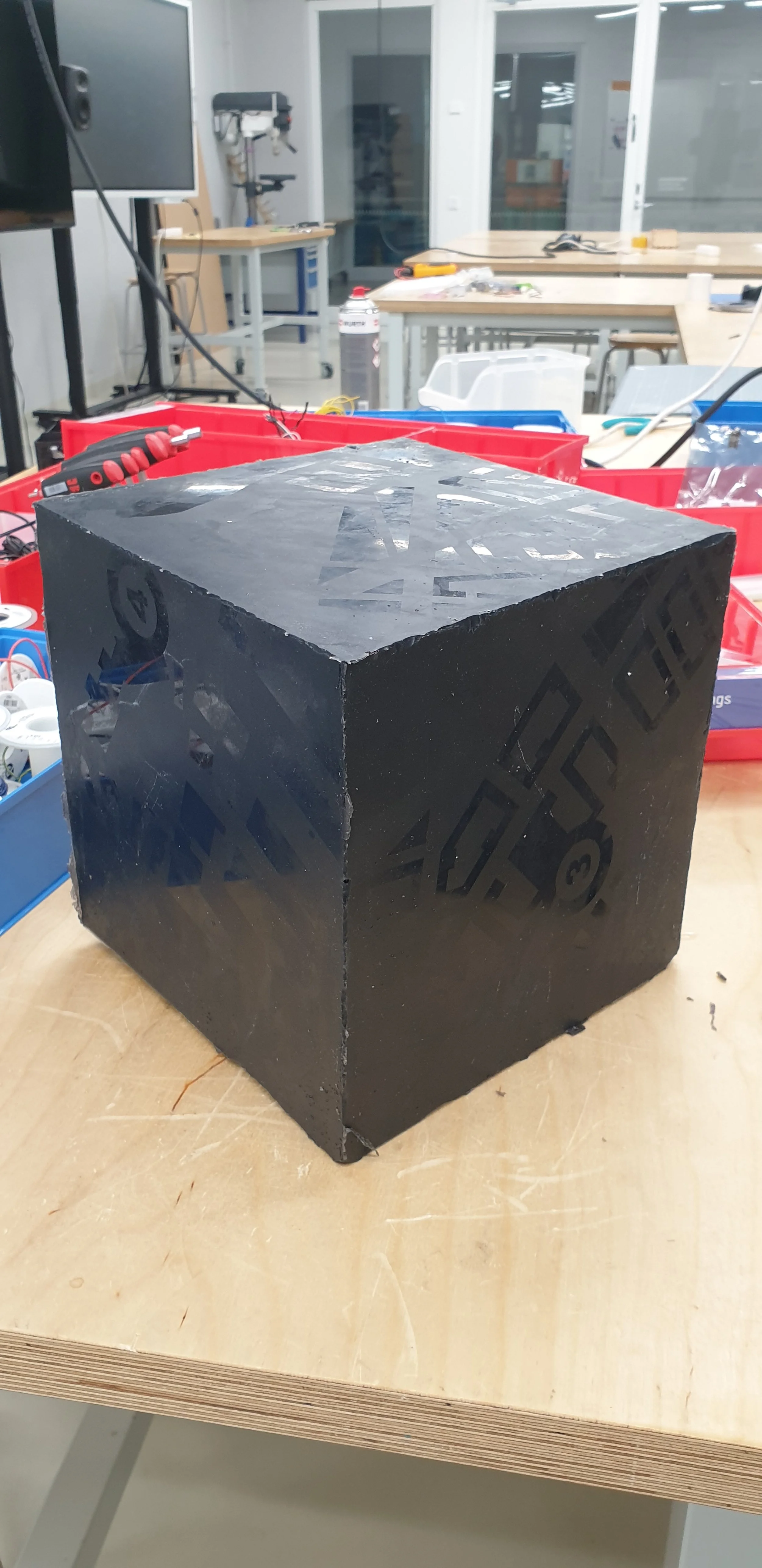 Mustavalkoinen laatikko, jossa on kyljessä geometrisiä kuvioita, asetettu puupöydälle työpajassa tai laboratoriossa.