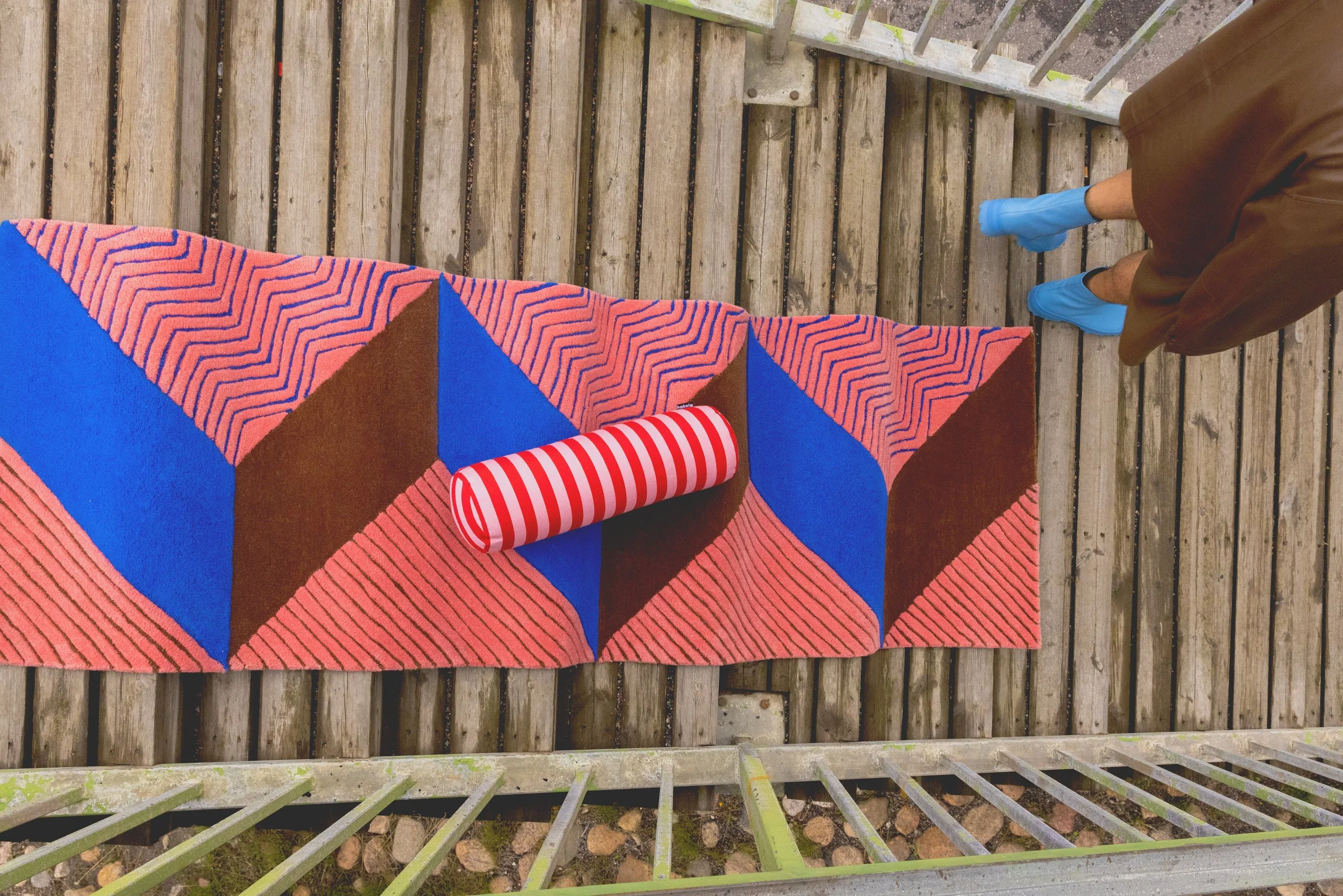 Kuvassa on värikäs matto, jonka yläosassa on kuvioita ja alaosassa tumma ja sininen väritys. Maton päällä on punavalkoraitainen paperi kääre. Kuvassa näkyy myös osa ihmisen jalkoja ja kenkiä sekä puista terassi ja metallikaiteet.