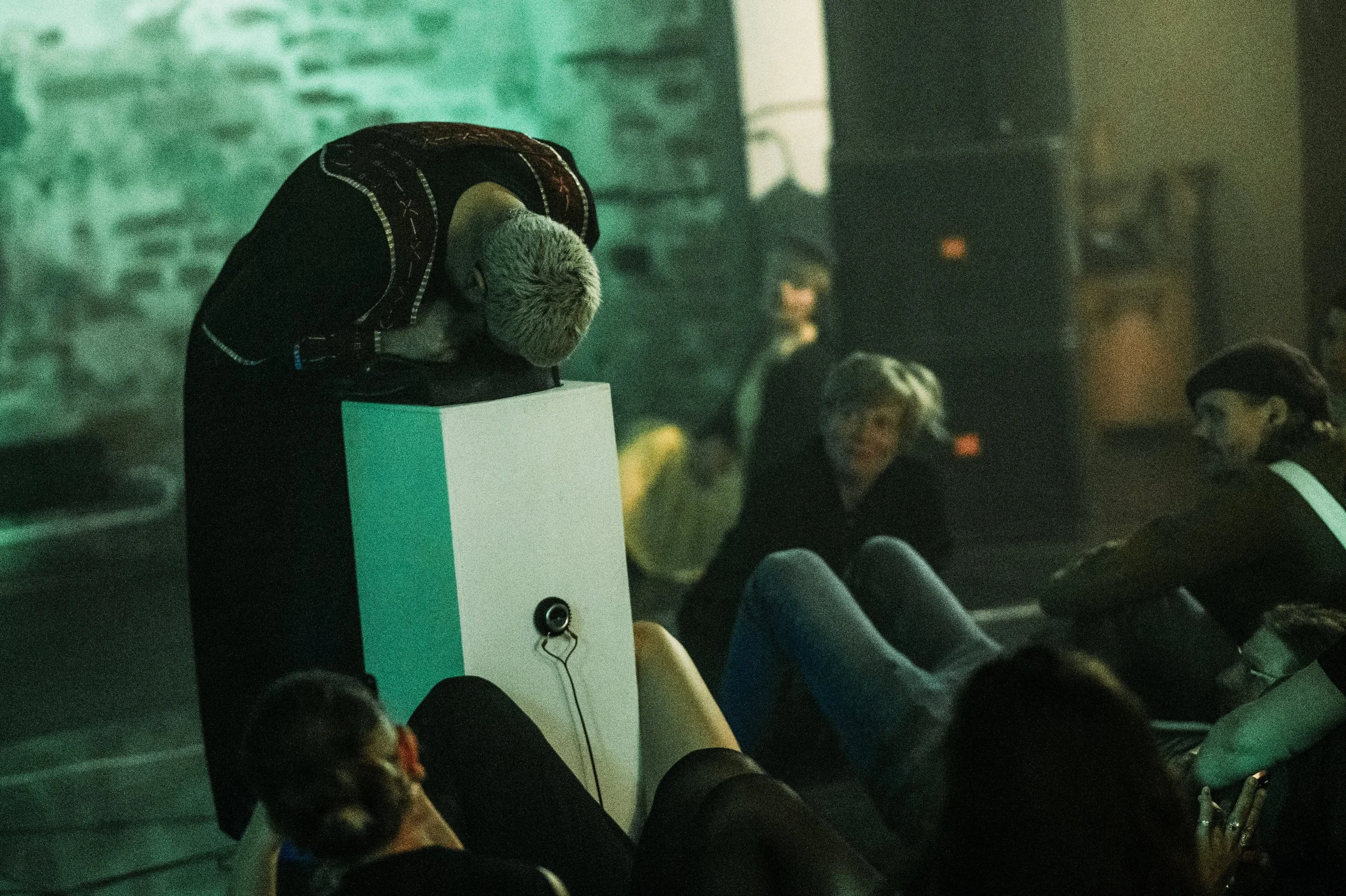 Henkilö nojaa ja kyykistyy kioskilla tai palkintopöydällä, muut ihmiset istuvat ja katsovat vaaleatukkaista henkilöä, viileän vihreä valaistus, sisätila.