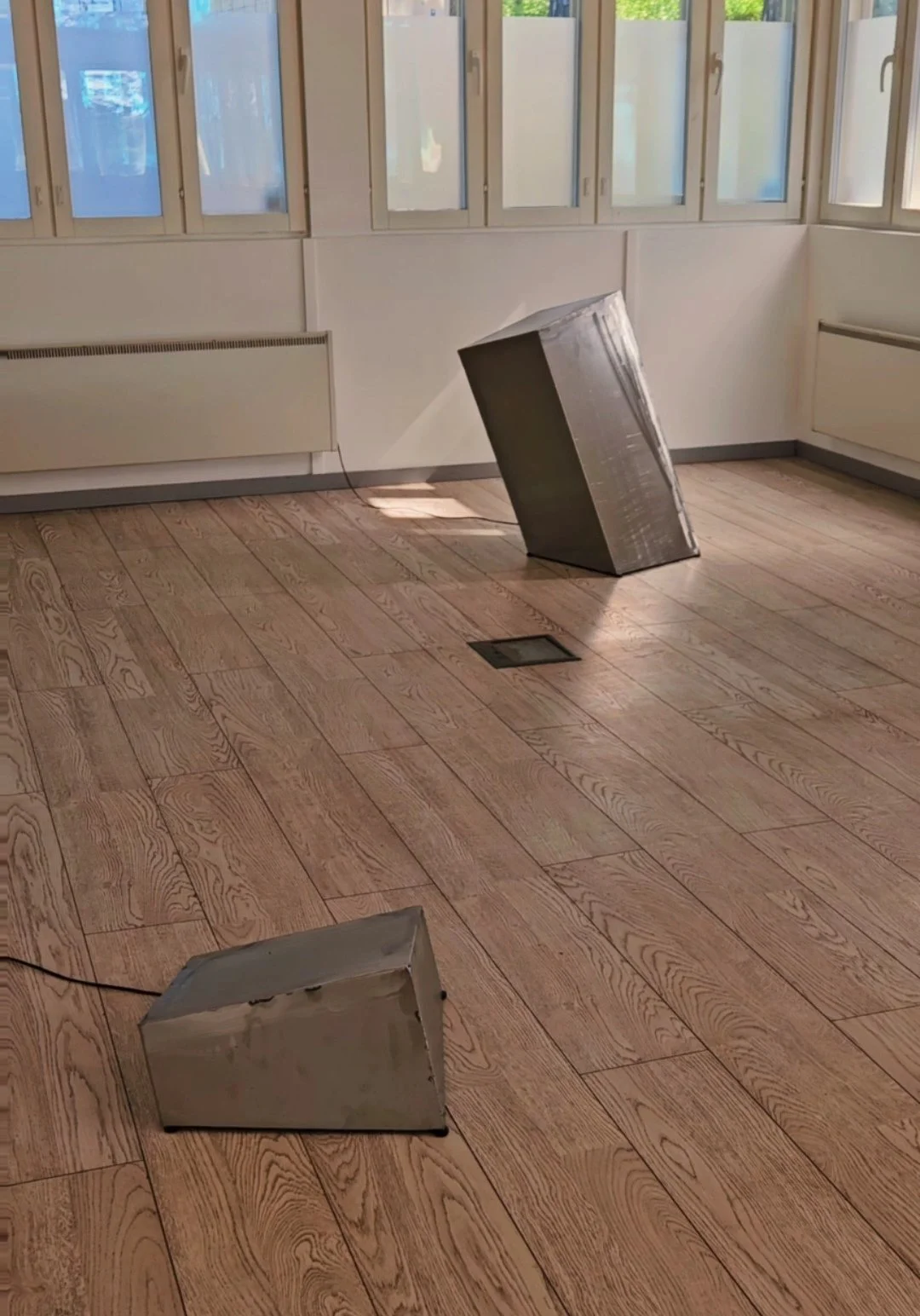 Kaksi metallihyllykköä, jotka ovat kaatuneet lattialle huoneessa, jossa on suuri ikkuna ja puukulautavisi. Lattialla on lattialämmitin ja kattolamppu.