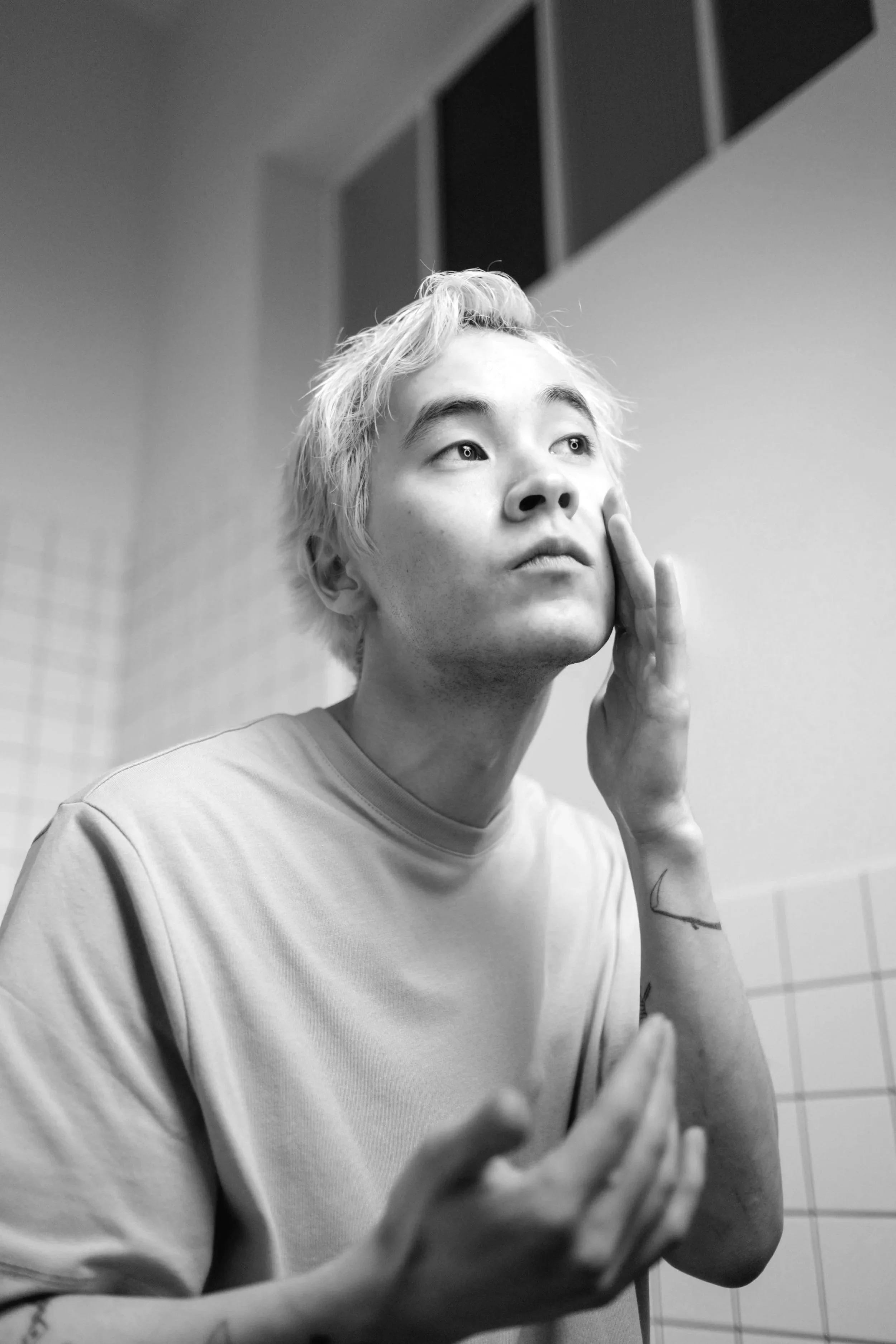 Black and white photo of a young man with light-colored, messy hair, touching his face gently with one hand and holding the other hand near his chest, standing in a room with tiled walls and a window.