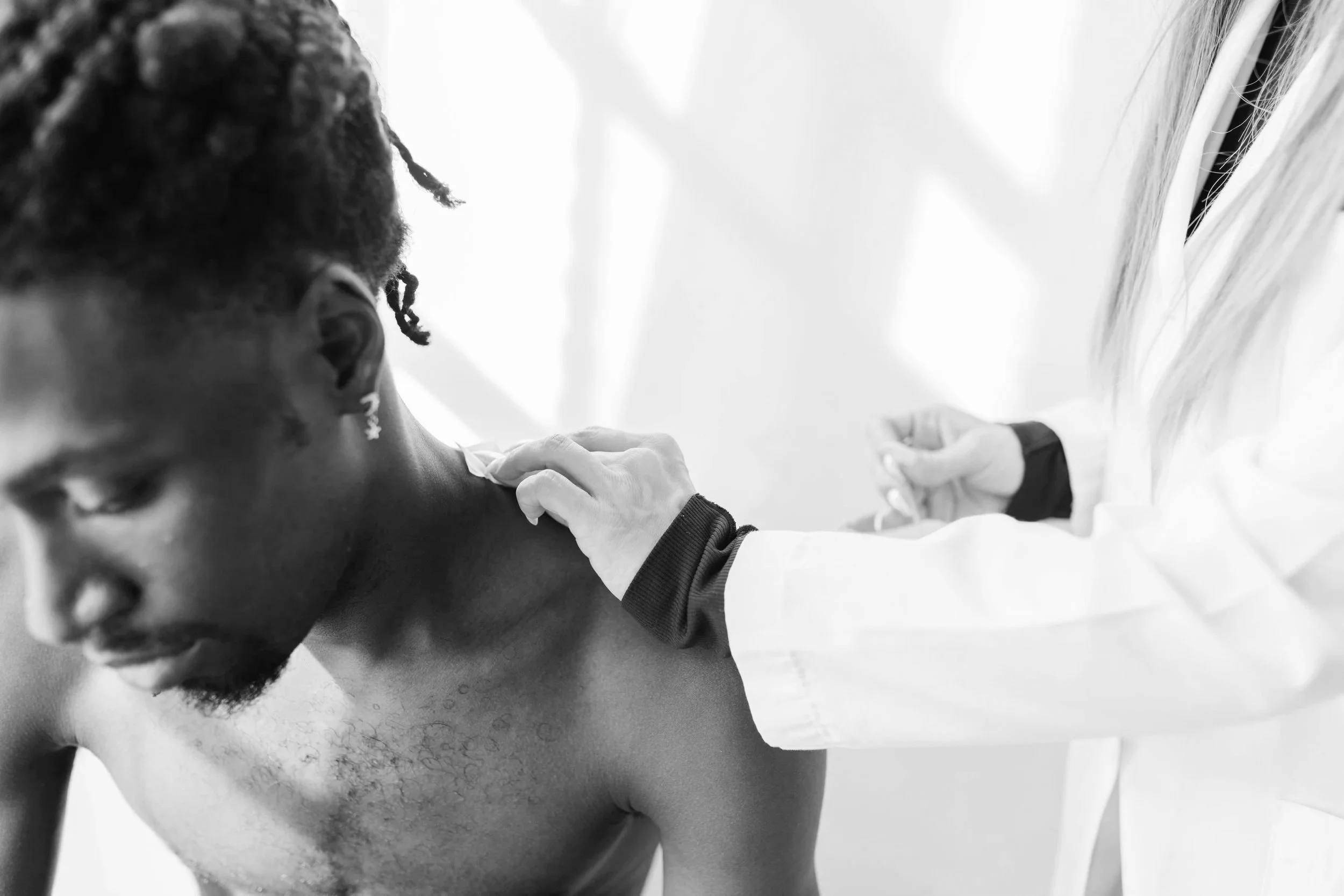 A healthcare professional administering a vaccine or an injection to a shirtless man with dark skin and textured hair in a clinical setting.