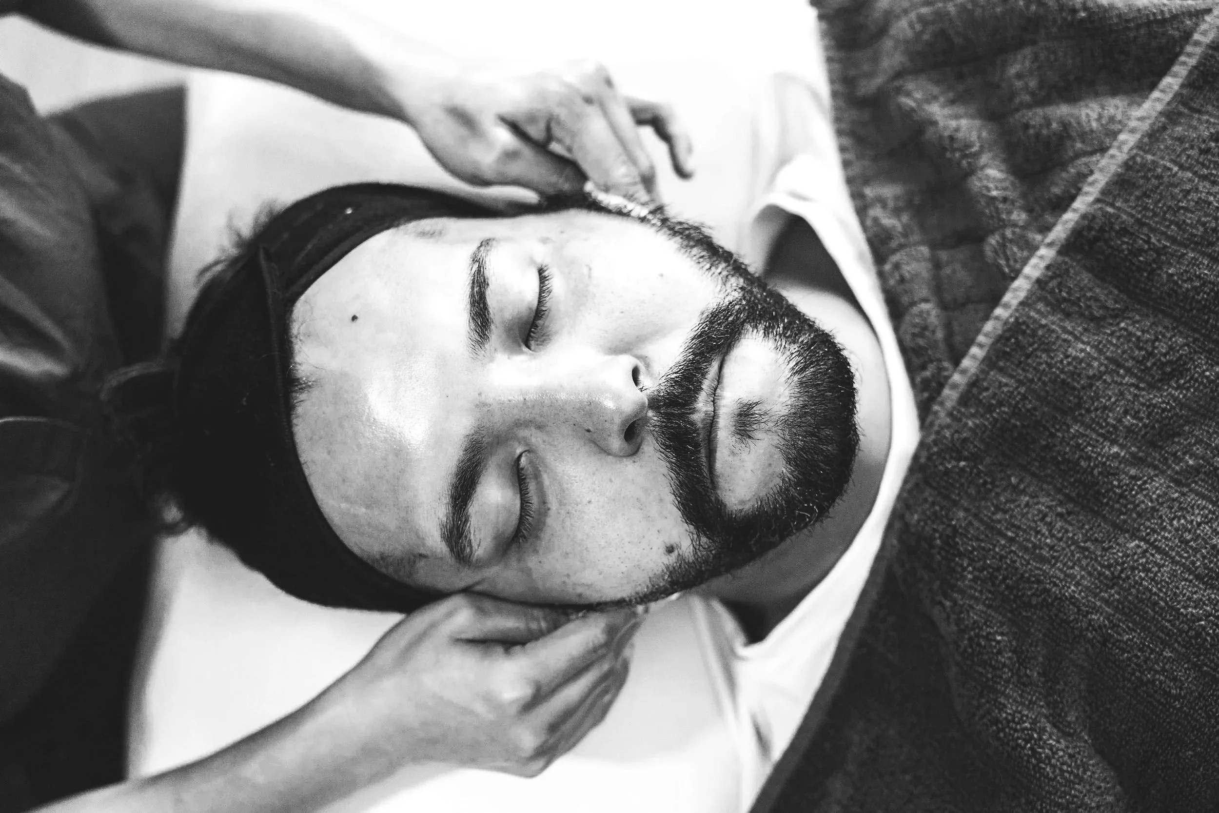 Black and white photo of a man sleeping with a beard and mustache, lying on his side, with hands gently touching his face.